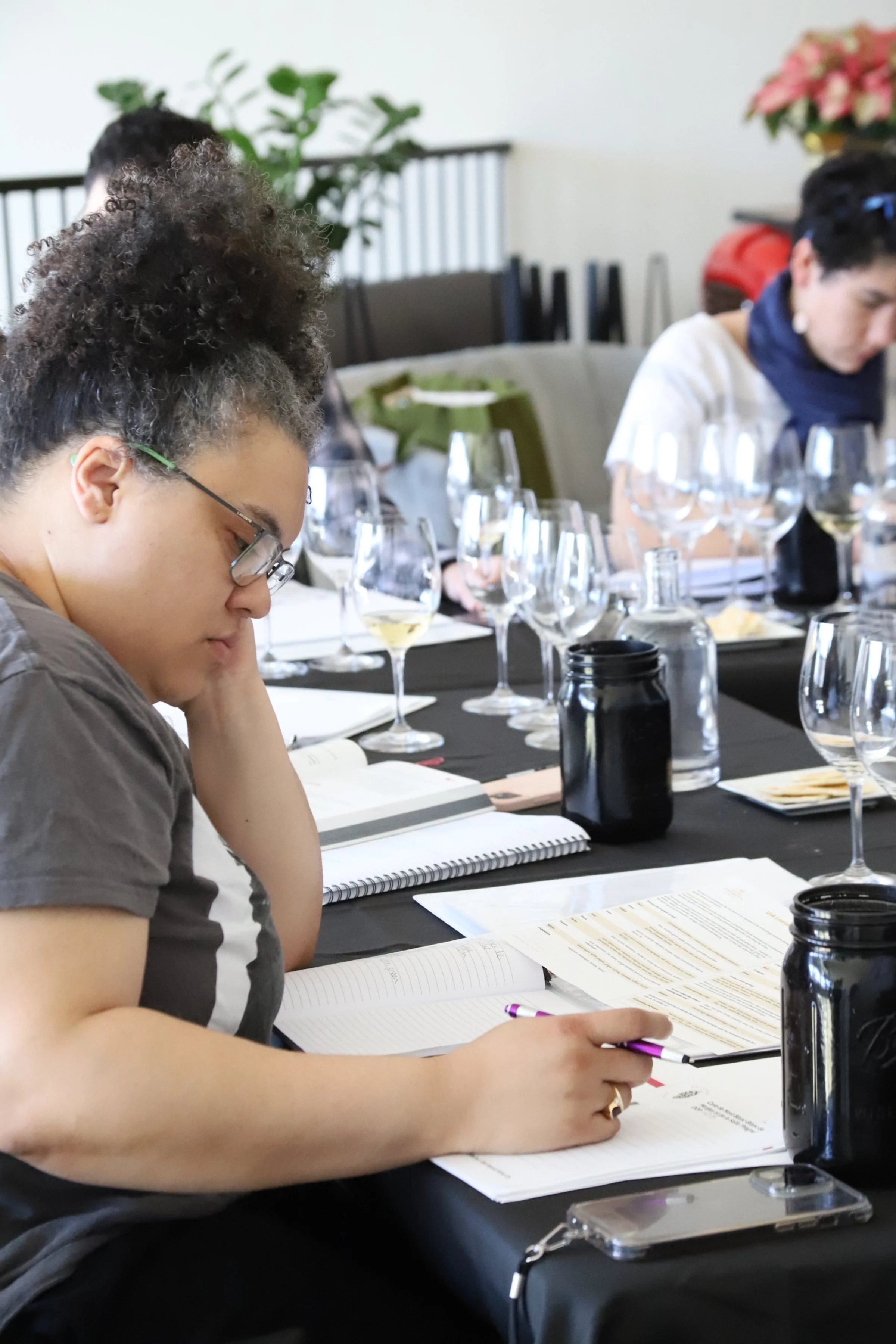 A woman with glasses and curly hair, taking notes at a conference or workshop, with multiple glasses of wine and documents on the table.