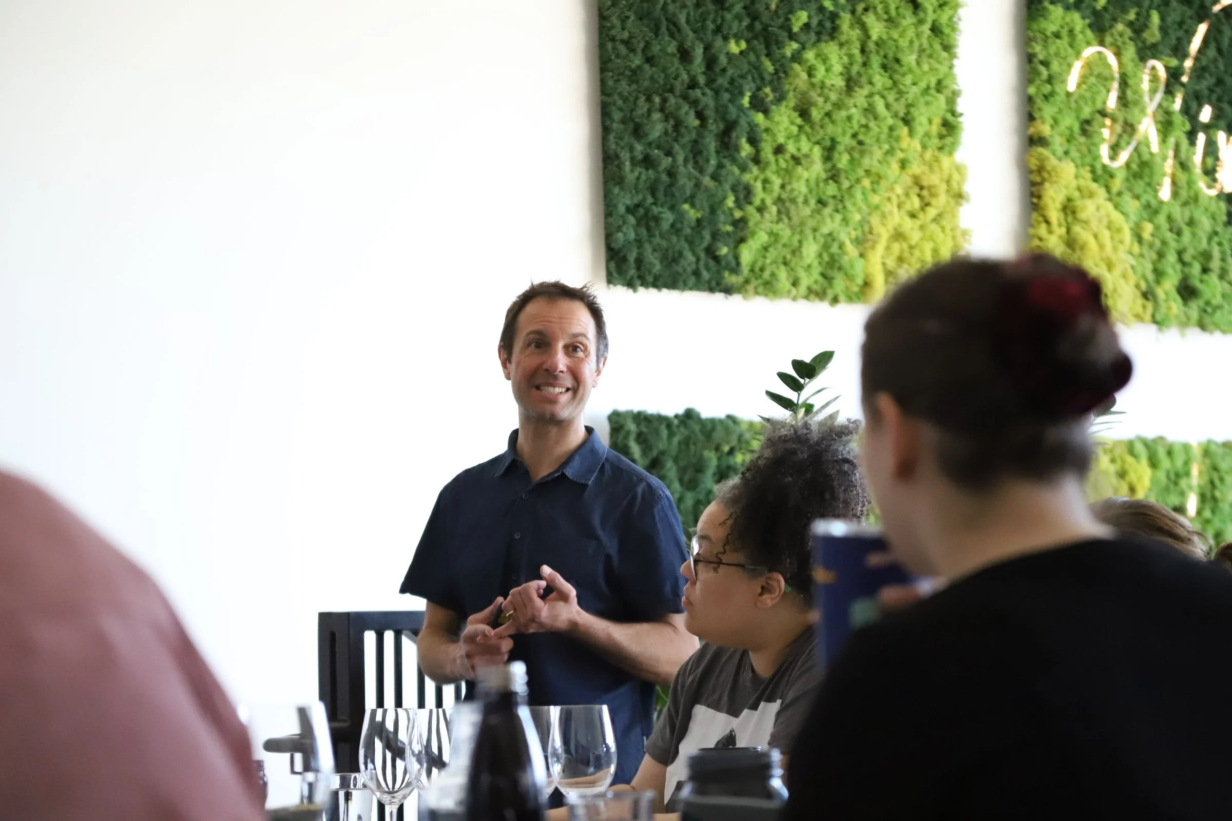 A man standing and smiling at a group of people seated at a table during a social gathering or meeting in a bright room with green wall art.