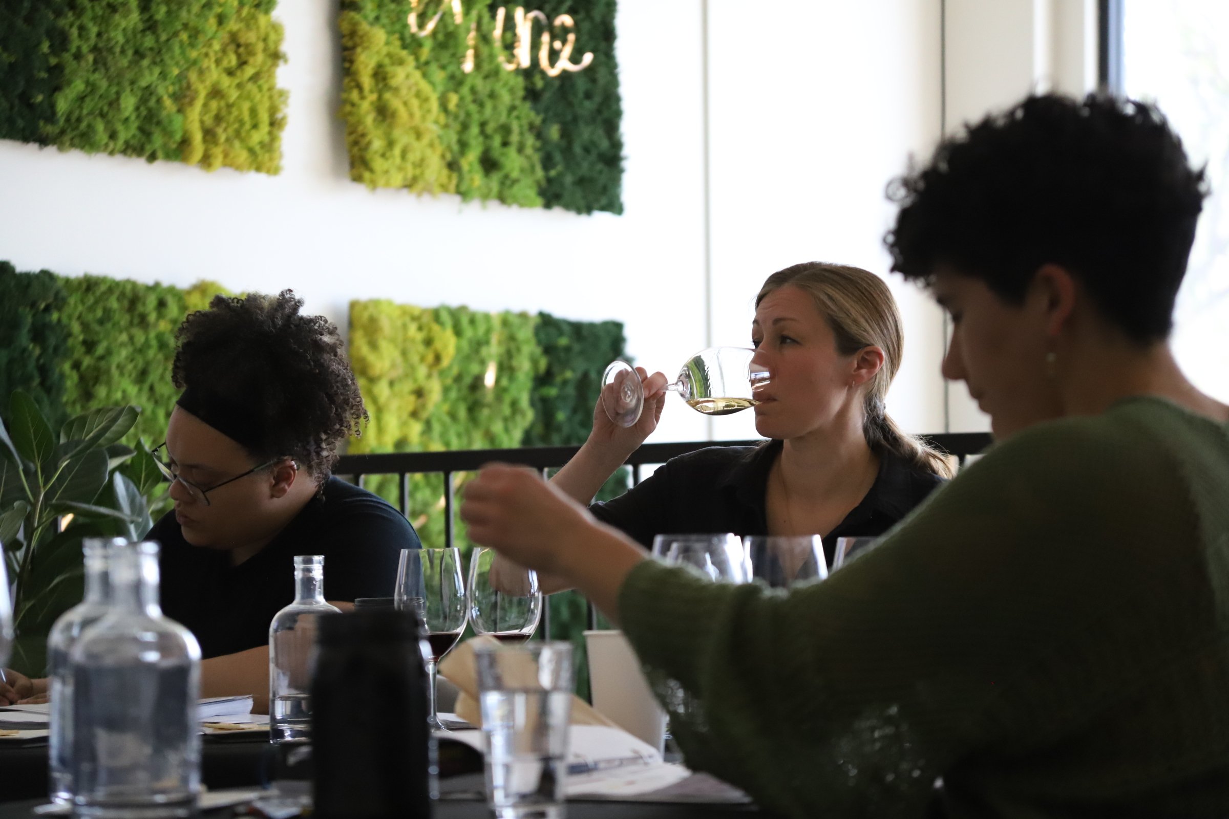 Three women seated at a table, one of them is drinking white wine, in a modern room with green moss wall art.