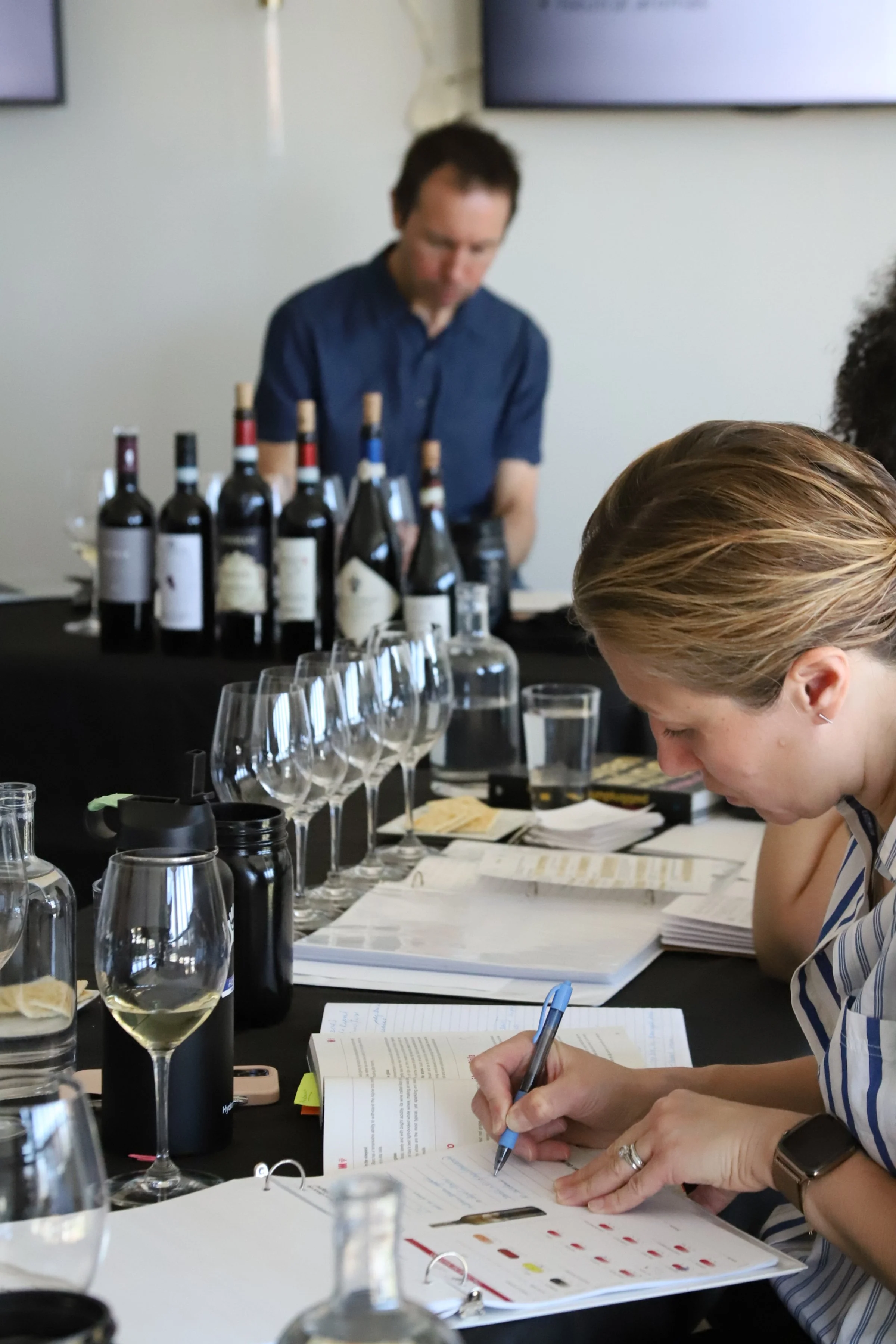 People participating in a wine tasting class, with bottles of wine and glasses on a table, and a instructor in the background.