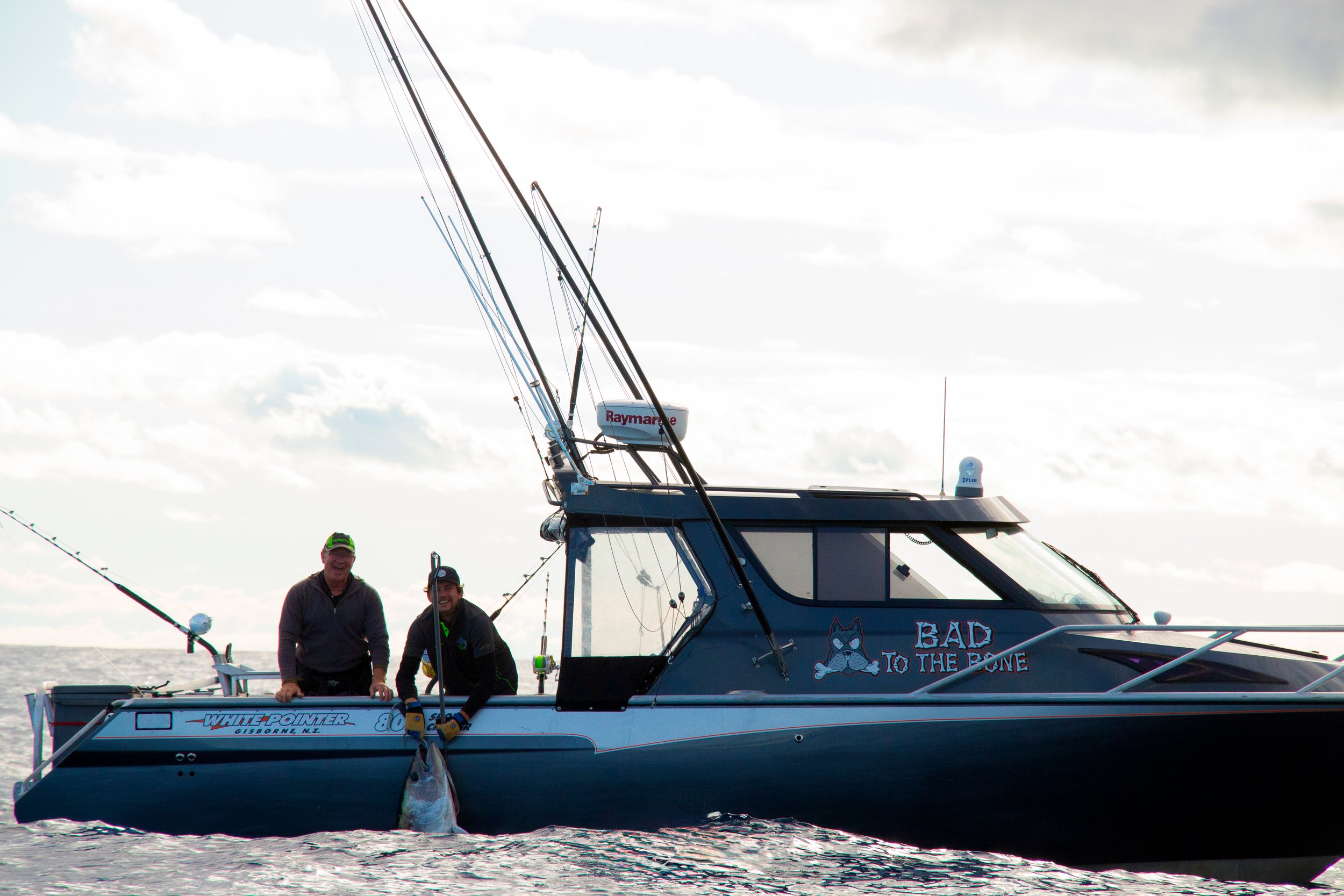 Two men on a fishing boat holding a large fish, with fishing gear and equipment, on a body of water under a cloudy sky.