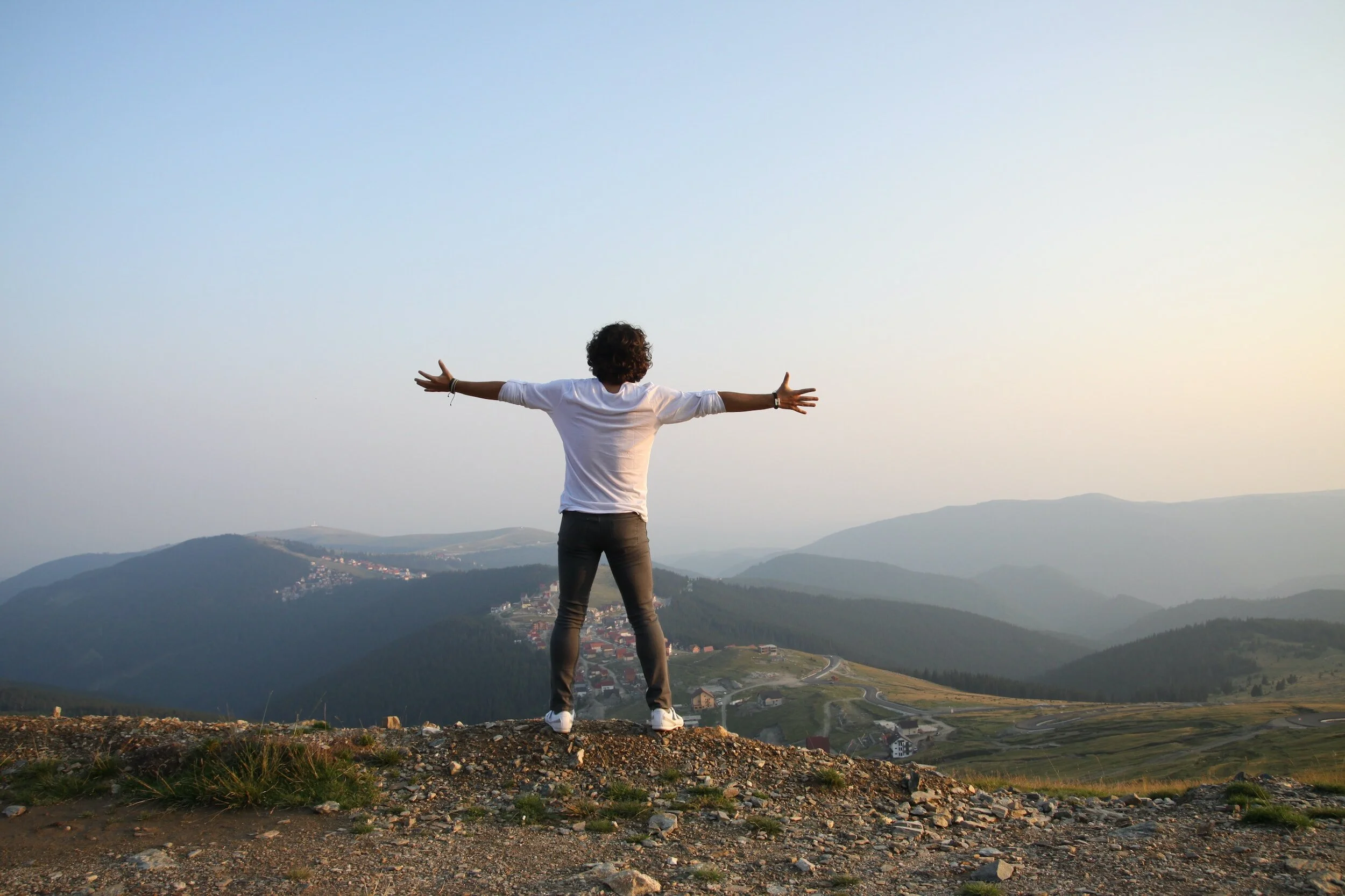 Person with arms outstretched, standing on a mountain overlooking a valley and distant hills.