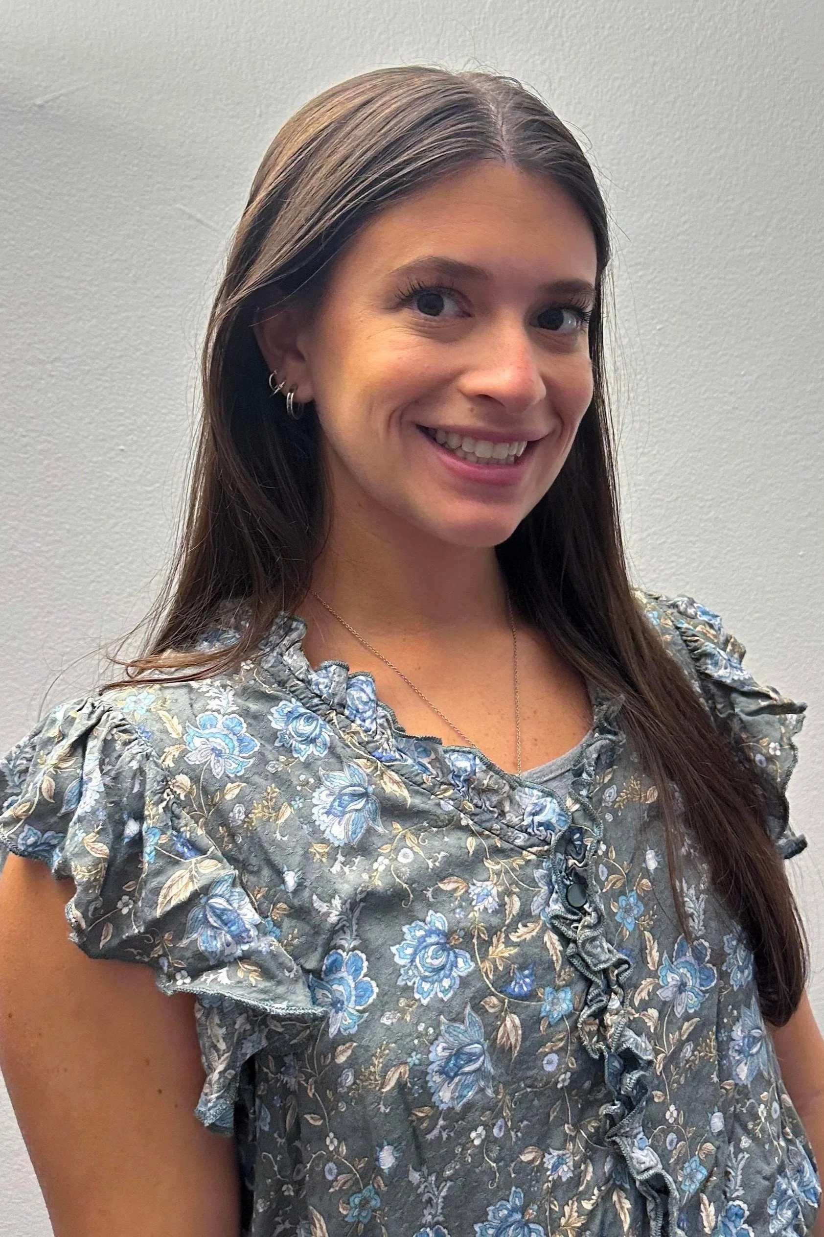 A smiling woman with long brown hair, wearing hoop earrings, a necklace, and a floral blouse, standing in front of a plain, light-colored wall.