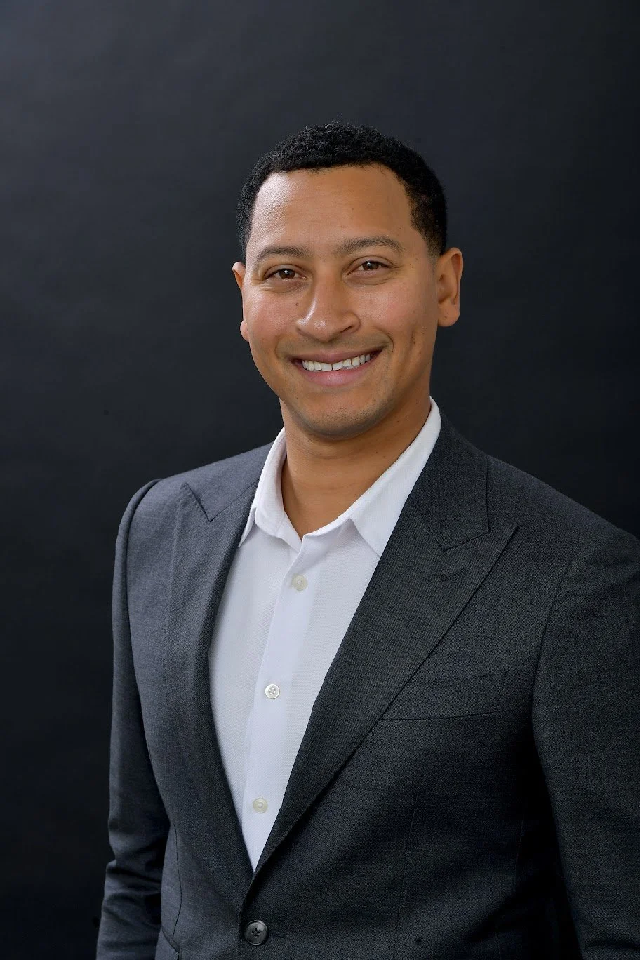 A smiling man in a dark gray suit and white shirt standing against a black background.
