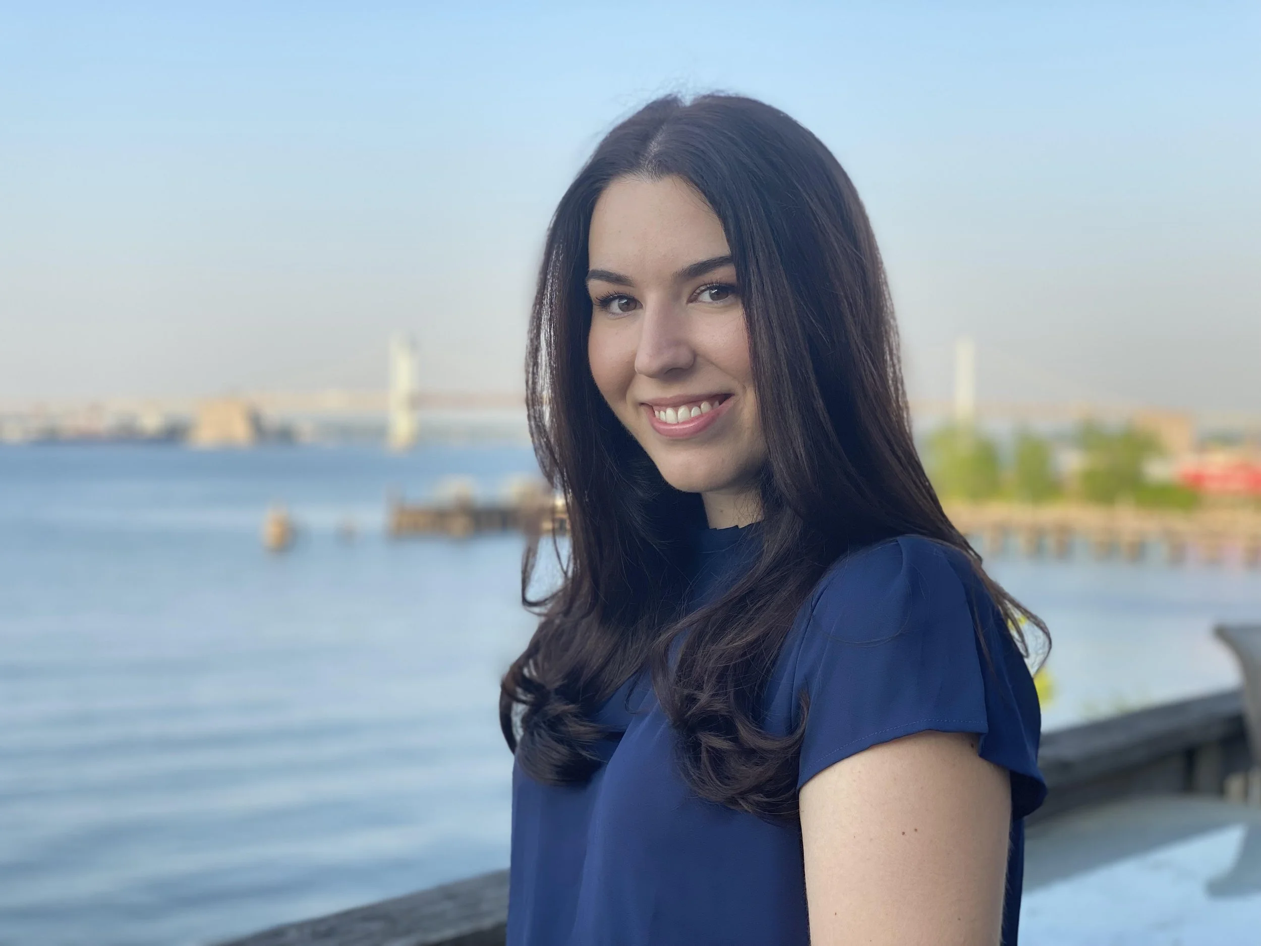 A young woman with dark brown hair smiling outdoors near a body of water with a bridge in the background.