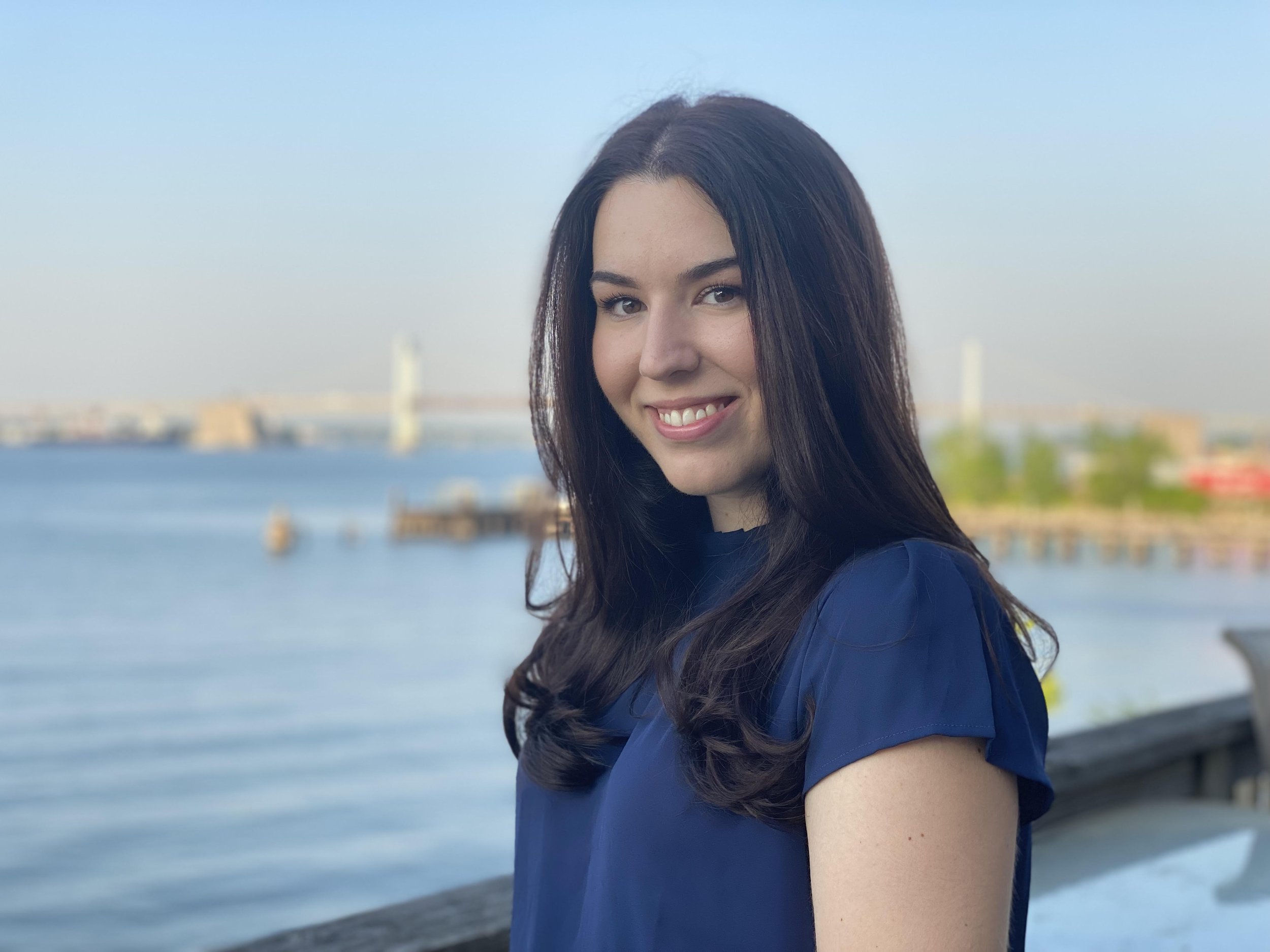 A smiling woman with long dark hair standing near a body of water with a bridge in the background.