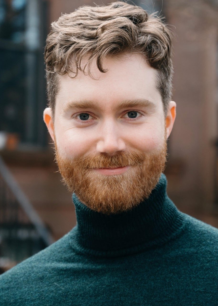 Close-up of a smiling man with blue eyes, curly light brown hair, and a full red beard, wearing a dark green turtleneck sweater.