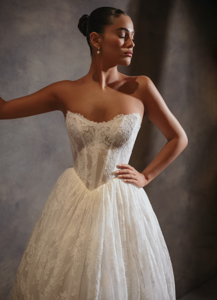 A woman in a white strapless lace wedding dress with her eyes closed and one hand on her hip, standing against a dark background.