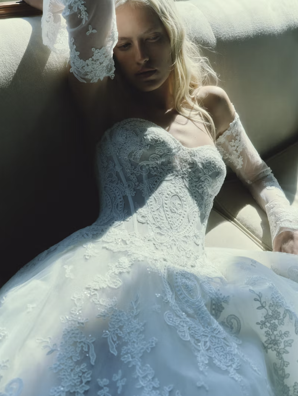 A woman in a white lace wedding dress sits on a couch, with sunlight casting shadows on her face and dress.