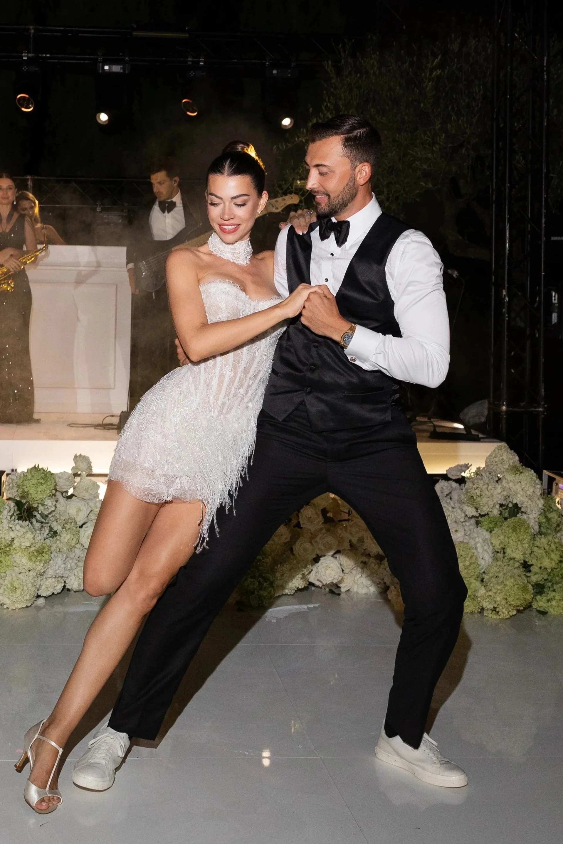 A man and woman dancing together at a formal event, with a band playing and floral decorations in the background.