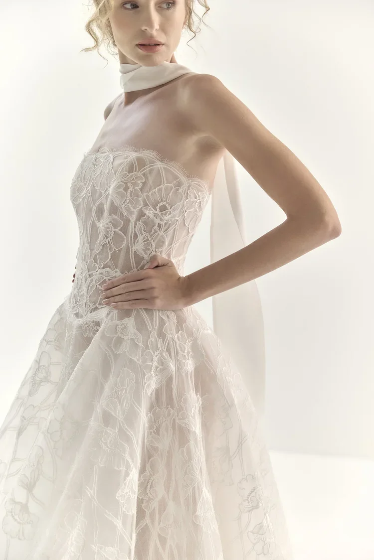 A woman wearing a strapless, floral lace wedding dress with a tulle skirt and a white neck scarf, posing with one hand on her hip against a plain white background.