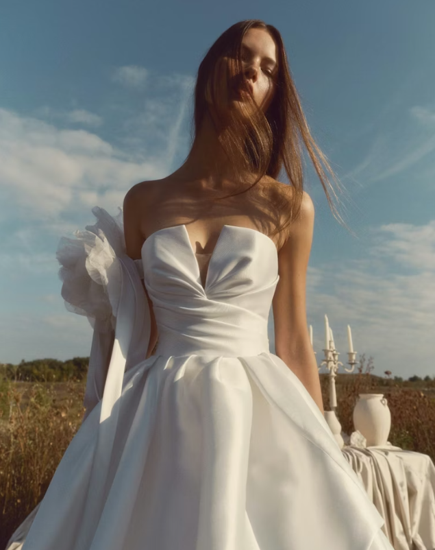A woman in a strapless white gown standing outdoors with a blue sky and minimal clouds, with a table of white decor items including candles and vases in the background.