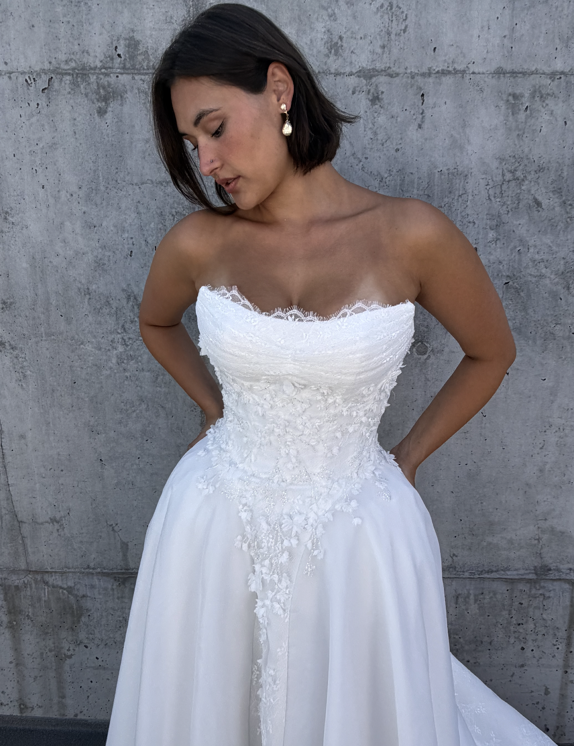 A woman wearing a white strapless wedding gown with lace details, standing against a gray concrete wall, looking down with her hands resting on her hips.