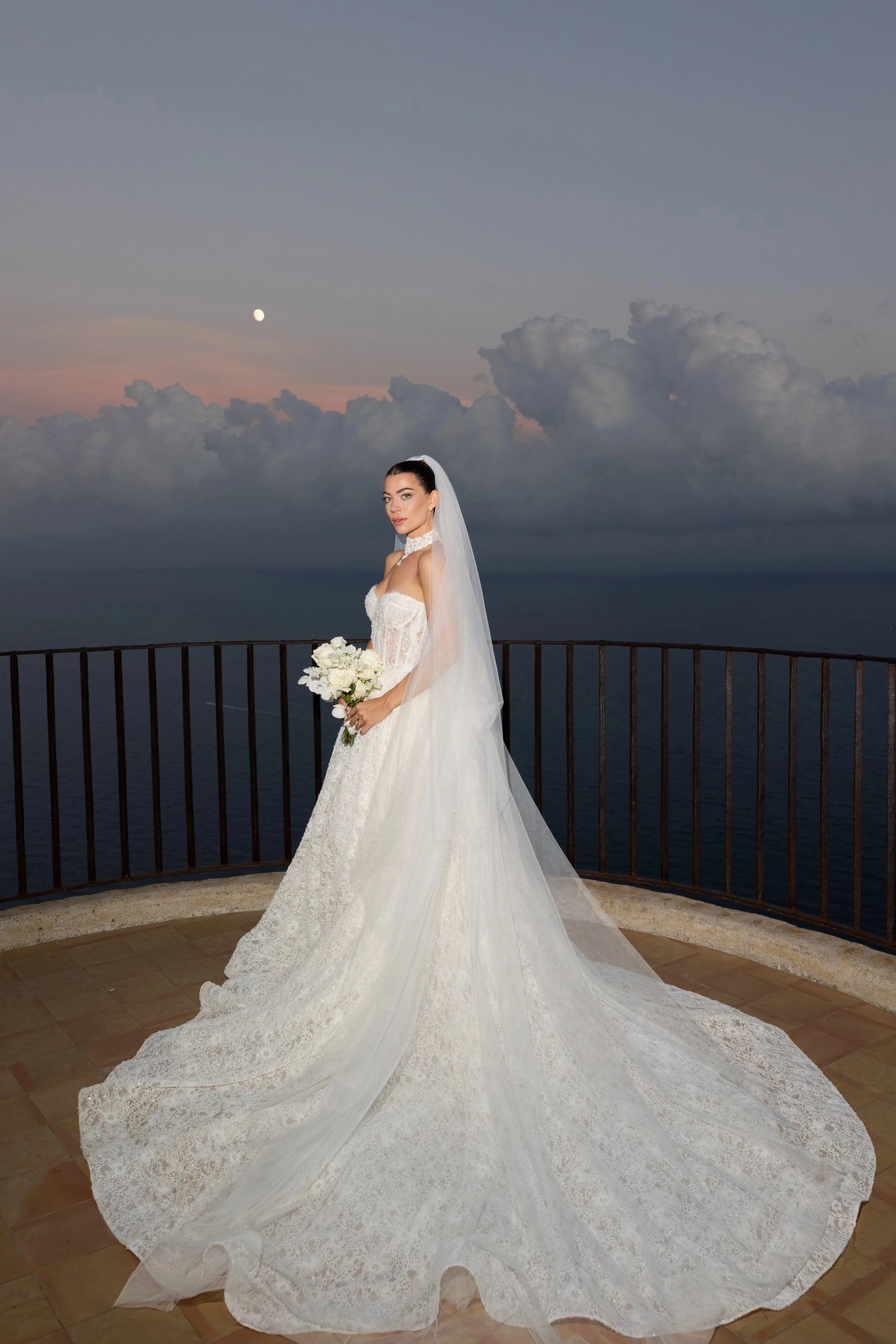 A bride in a white wedding gown holding a bouquet of white flowers. She stands on a balcony with a railing overlooking the ocean. The sky is partly cloudy with a visible moon, and it appears to be around sunset or sunrise.
