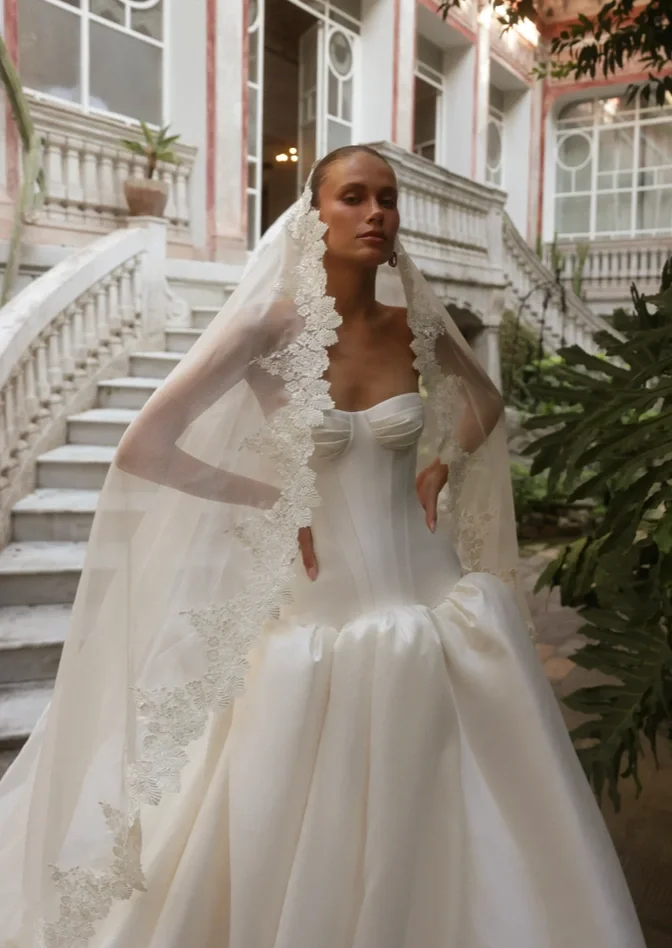 A woman wearing a vintage white wedding gown with lace trim and a veil, posing on a staircase outside a building with classical architecture and potted plants.
