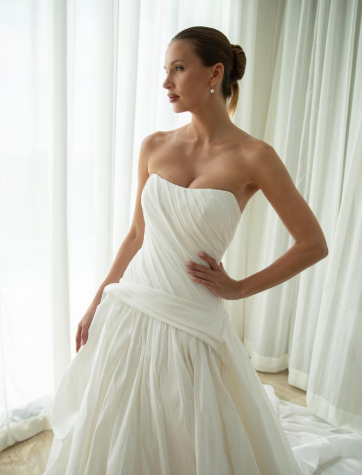 A woman in a strapless white wedding gown standing near sheer curtains.