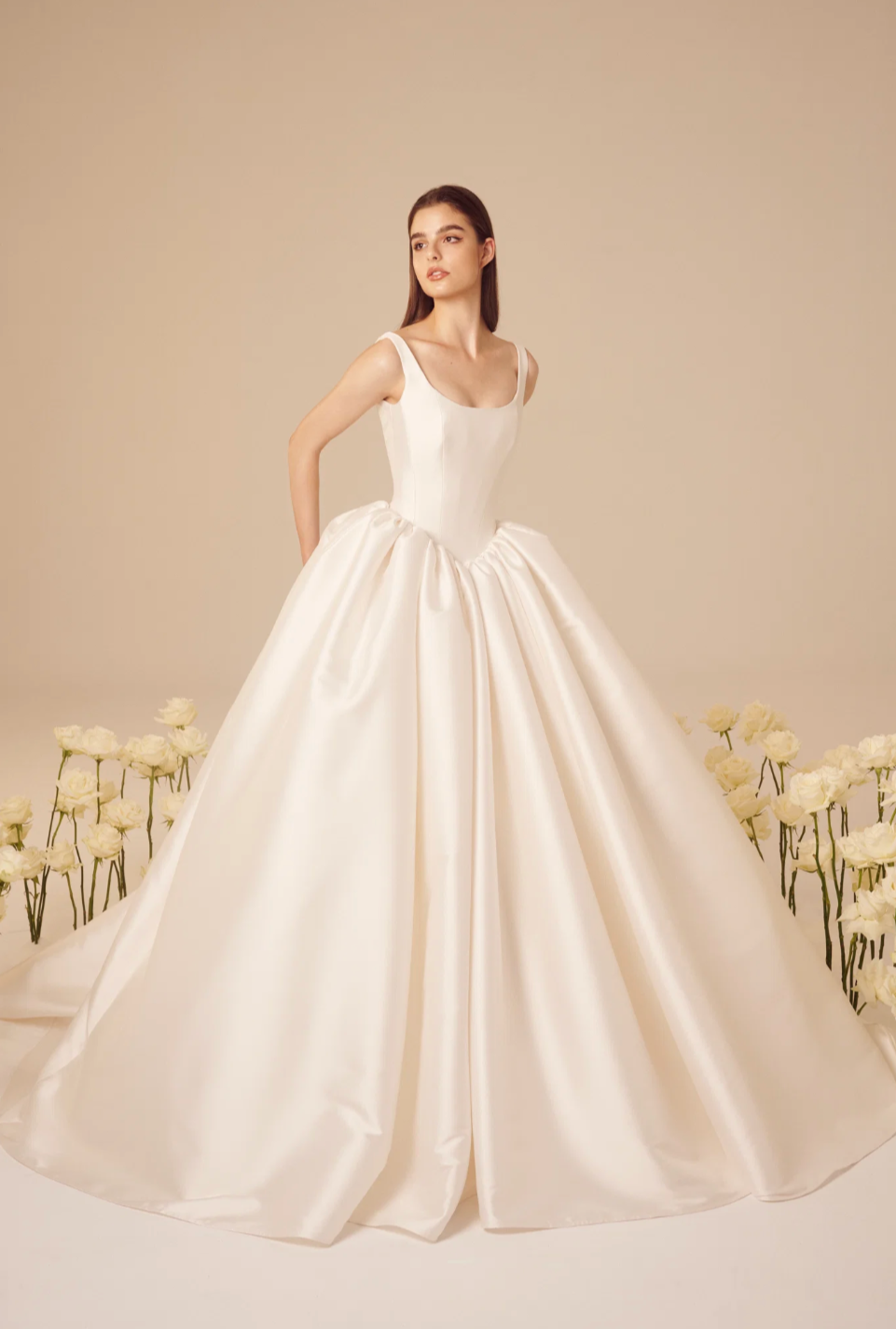 Young woman in a white wedding dress, standing among white flowers against a beige background.