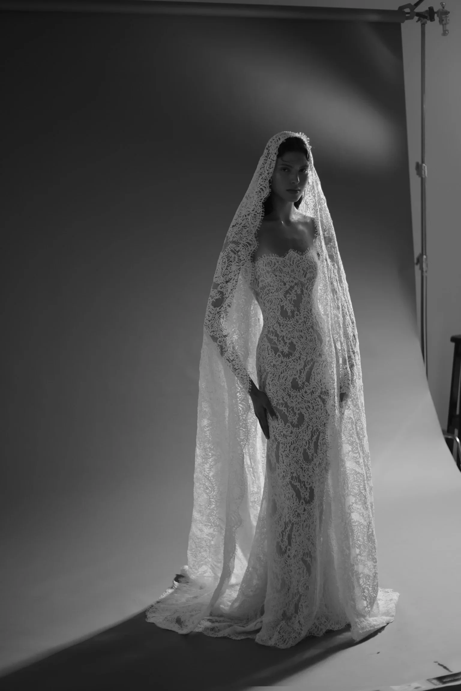 A woman wearing a lace wedding gown with a matching lace veil stands in a professional photography studio with a dark backdrop.