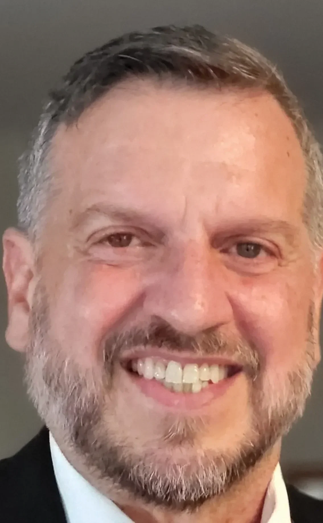 Close-up photo of a middle-aged man with short hair, a beard, and a wide smile showing his teeth, wearing a black suit and white shirt.