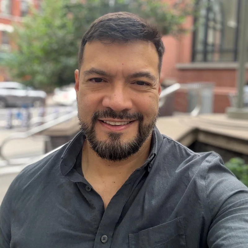 A man taking a selfie outdoors on a city street with trees and brick buildings in the background.