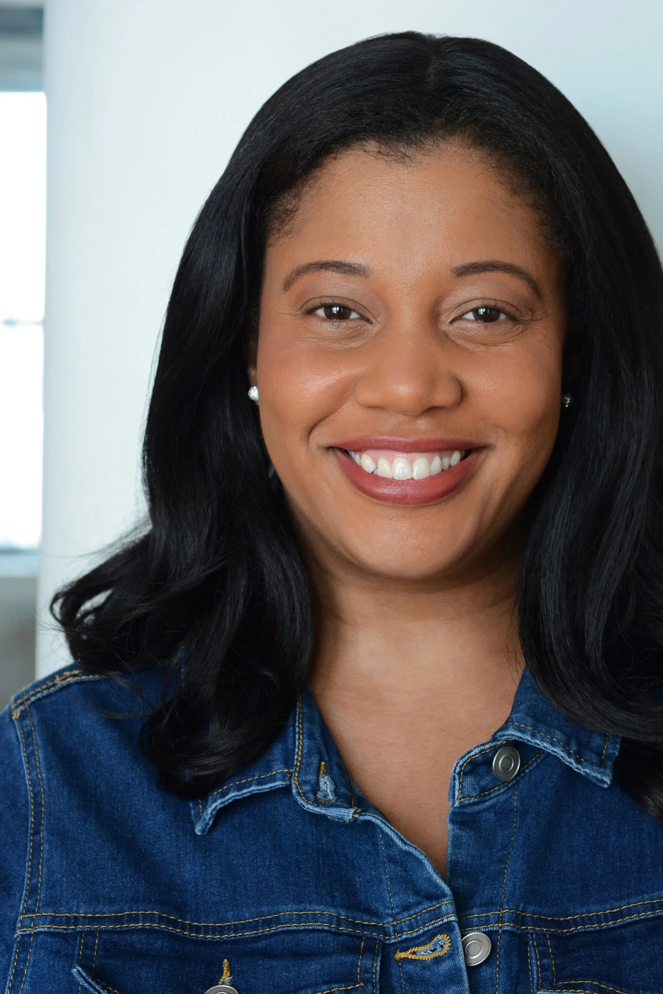 A woman with long black hair smiling, wearing a blue denim jacket and pearl earrings, standing indoors in front of a white wall with windows.