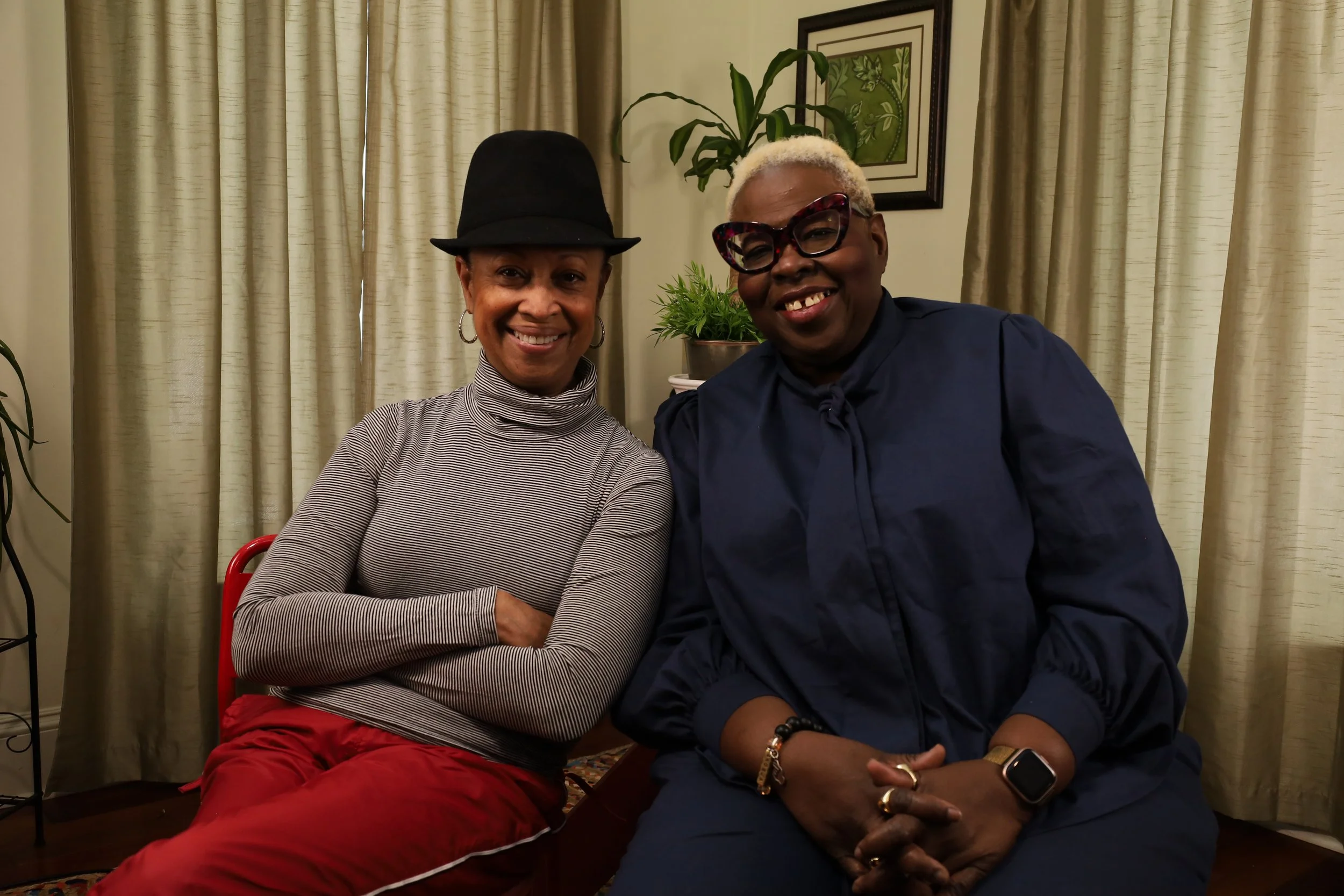 Two women sitting together indoors, smiling at the camera. One wears a black hat and red pants, the other has short blonde hair with glasses and dark blue clothing.