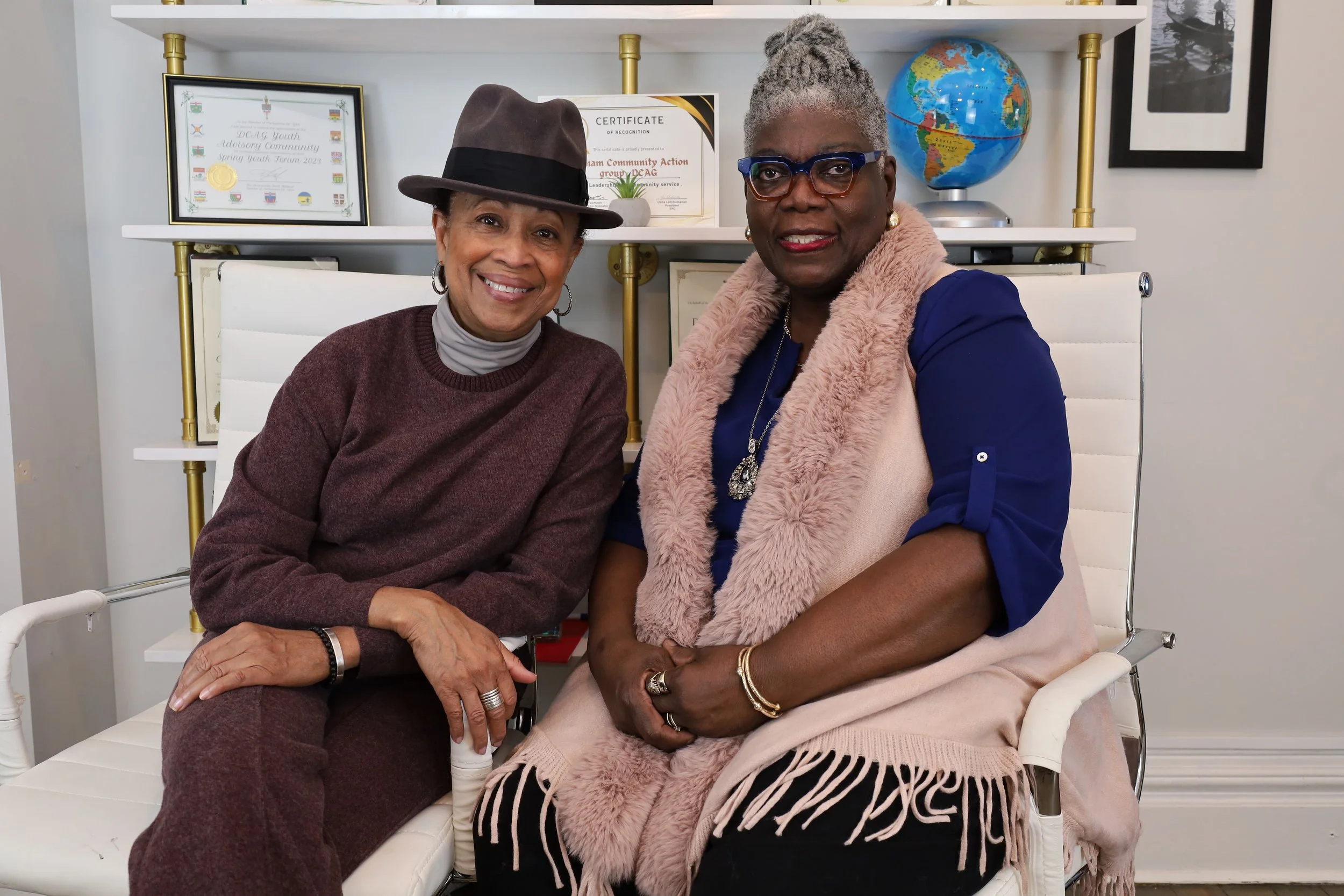 Two women sitting on white chairs in an office, smiling at the camera. The woman on the left is wearing a brown hat, a gray turtleneck, and a brown sweater. The woman on the right is wearing glasses, a blue blouse, and a pink fur vest. Behind them is a white bookshelf with framed awards, a certificate, a globe, and a black-and-white photograph.