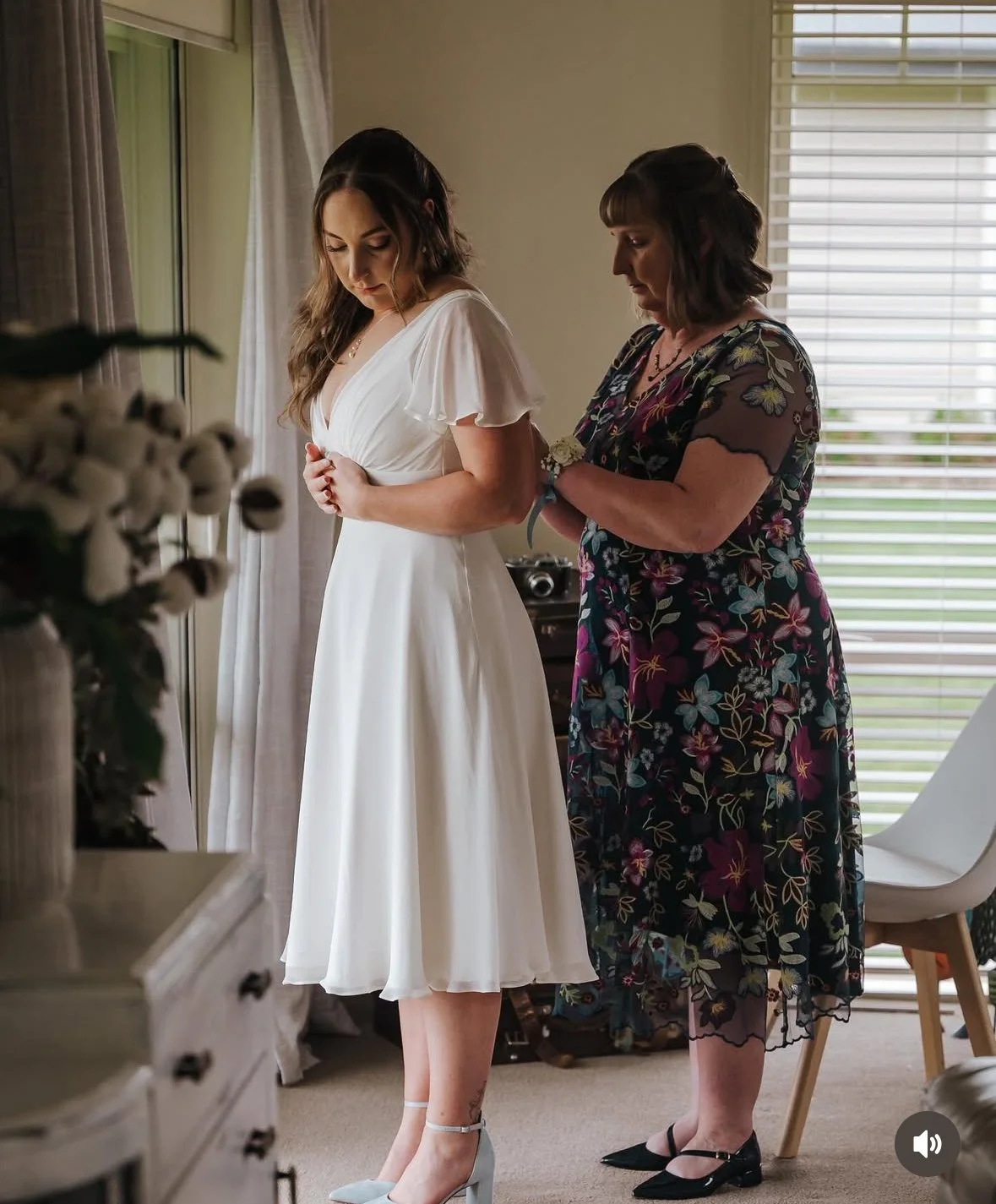 A bride in a white dress is being helped into her dress by a woman in a floral dress inside a well-lit room with large windows.