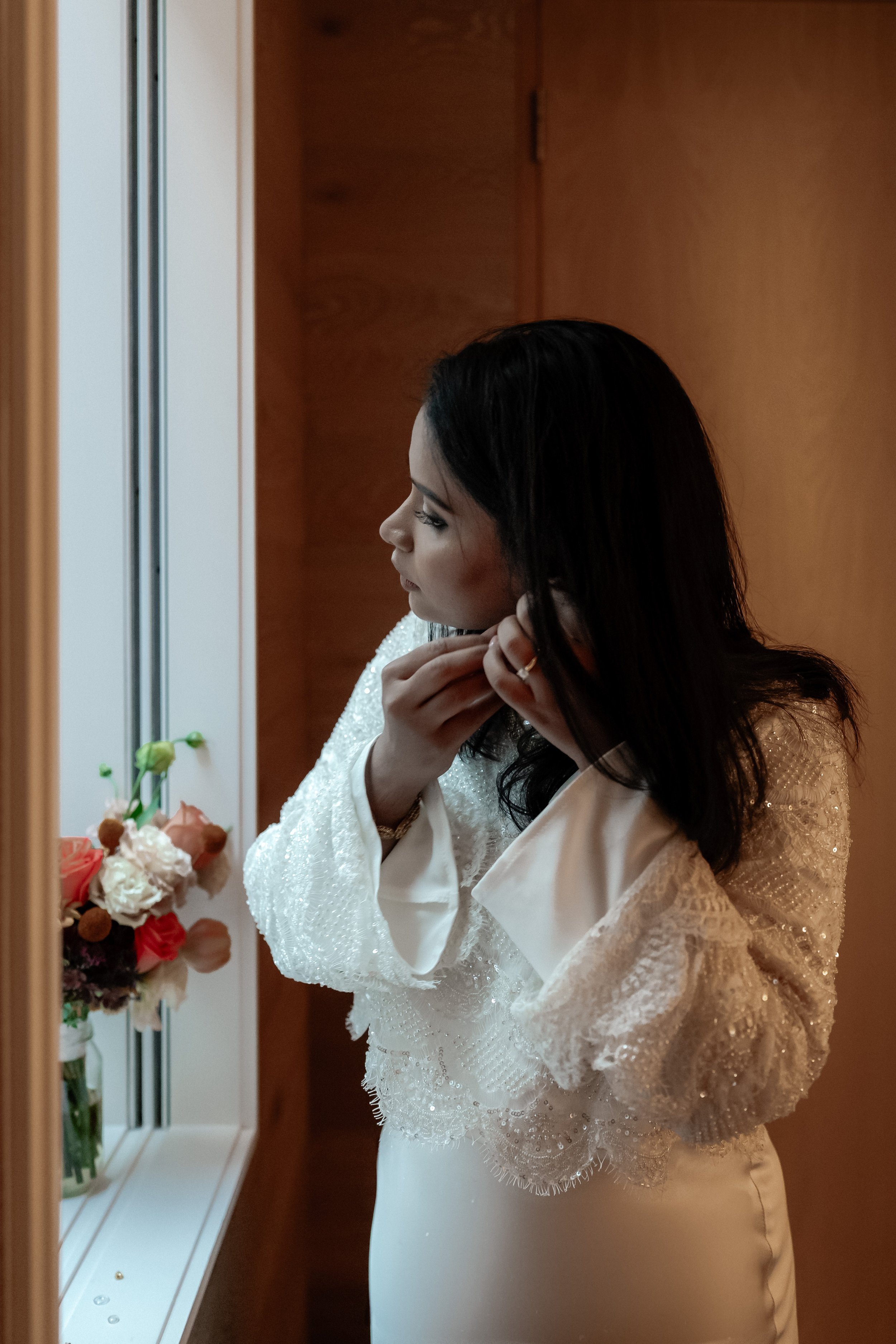 A bride getting ready in the mirror, putting on earrings.  She is wearing a sequined top