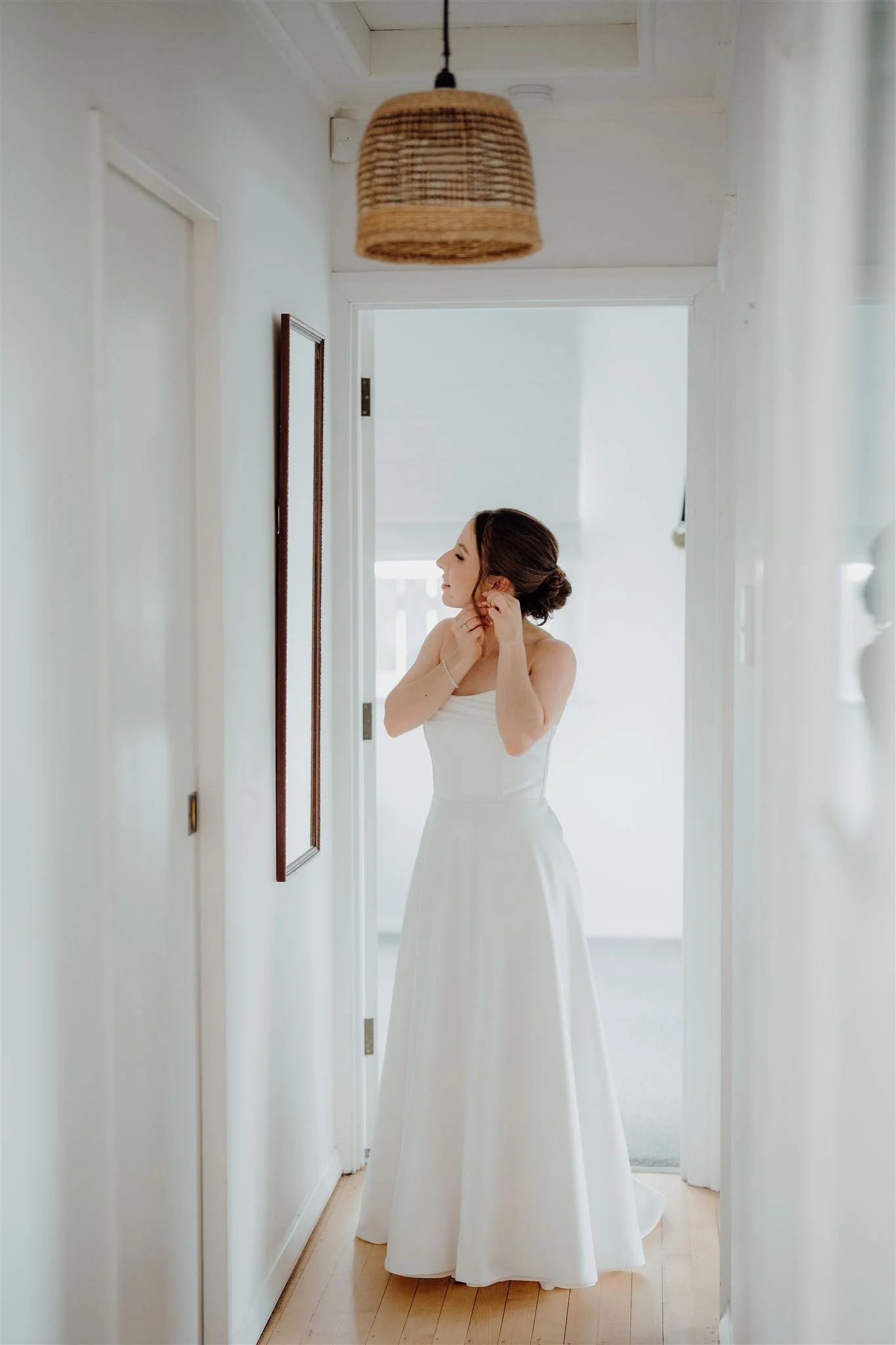 A woman in a white dress standing in a hallway, adjusting her earrings.