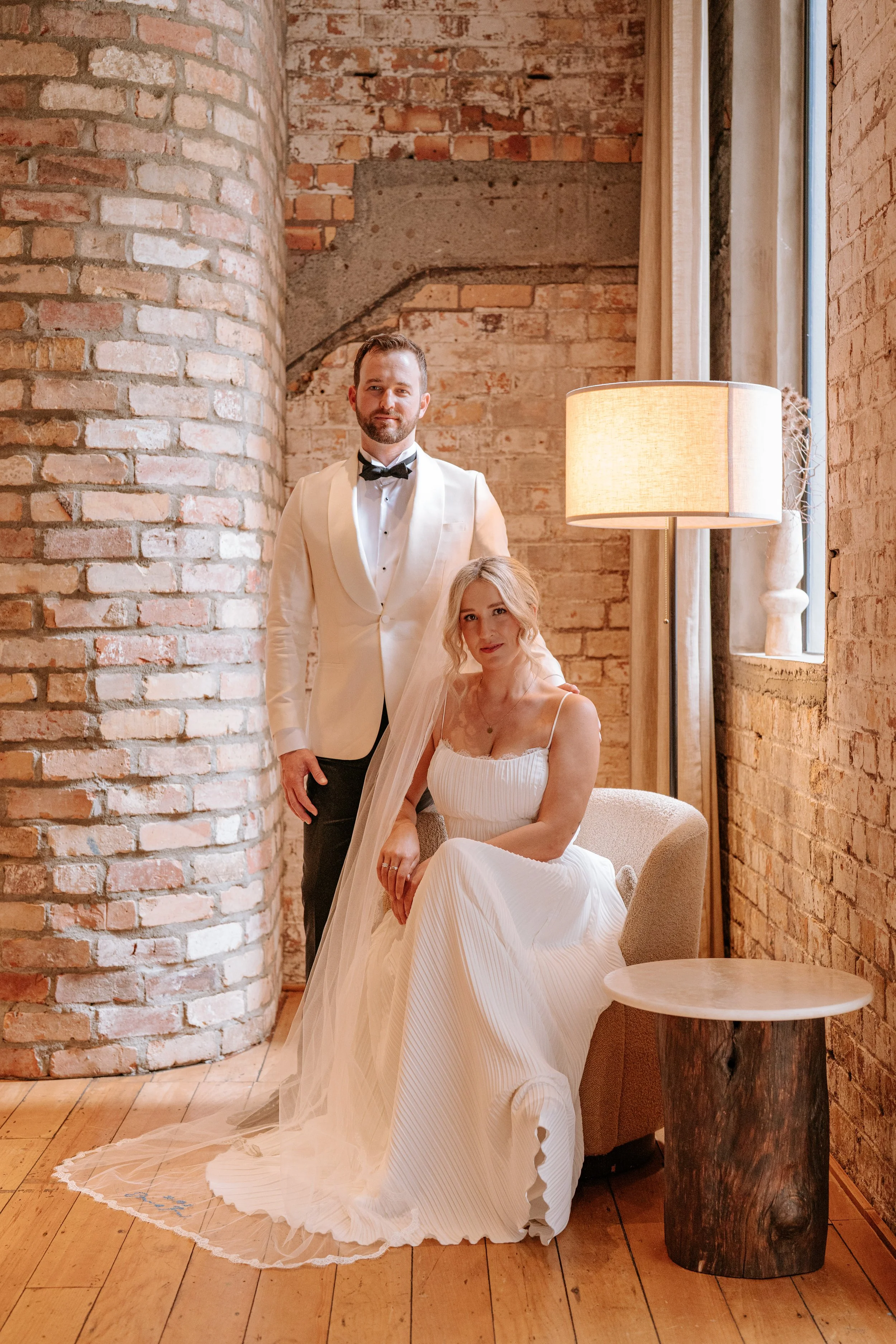 A bride in a white dress with thin straps and a groom in a white tuxedo with black bowtie posing in a warmly lit room with exposed brick walls, wooden floors, a window, and furniture including a beige armchair and a small wooden side table.