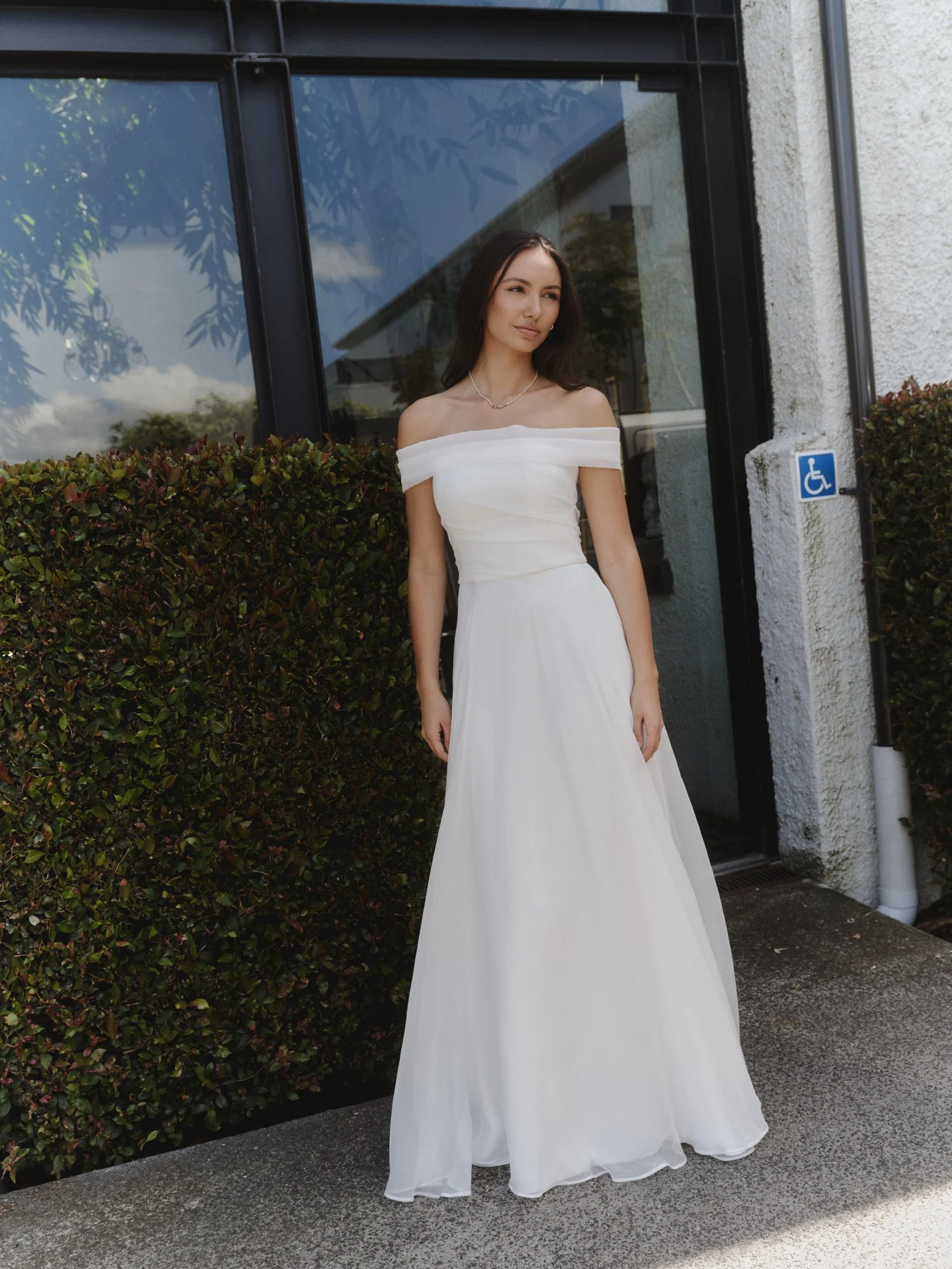A woman in a white off-shoulder dress standing outside near a glass building with a hedge and a handicap parking sign.