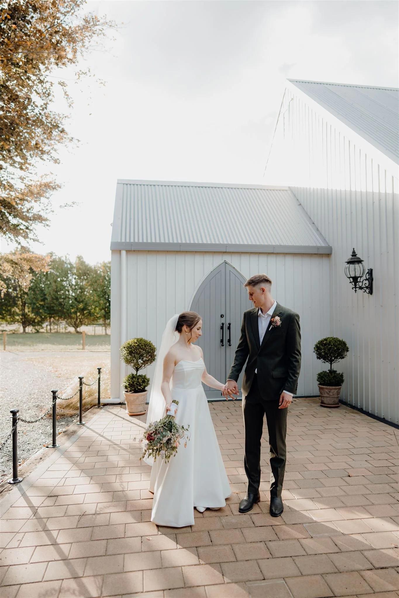 Bride and groom holding hands outside a white chapel, with a clear sky and trees in the background.
