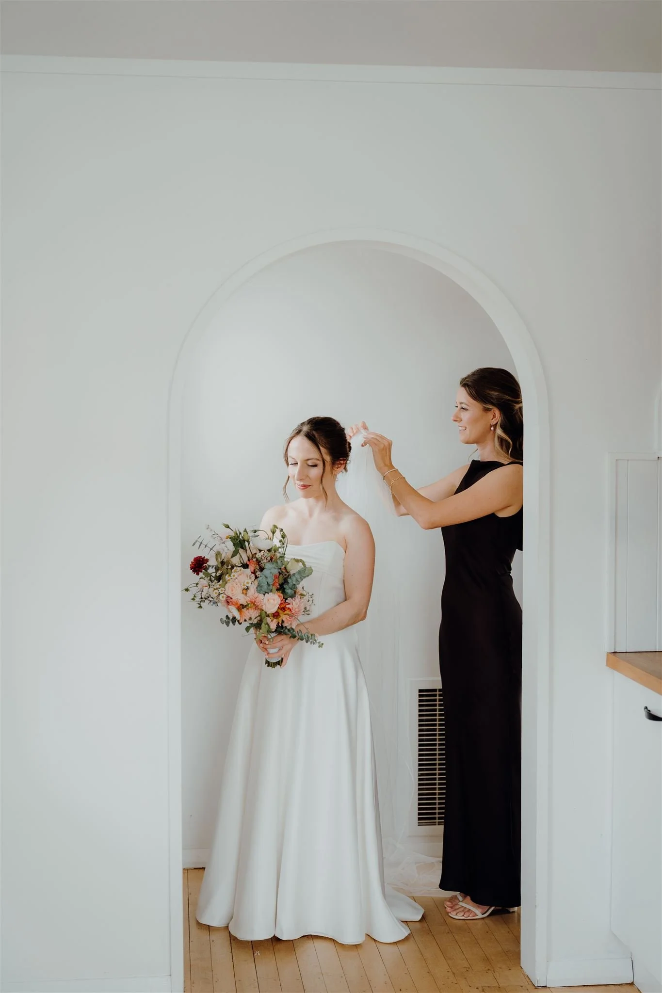 A bride in a white wedding dress holding a bouquet of flowers standing next to a woman in a black dress who is adjusting the bride's veil inside a small, white, arched alcove.