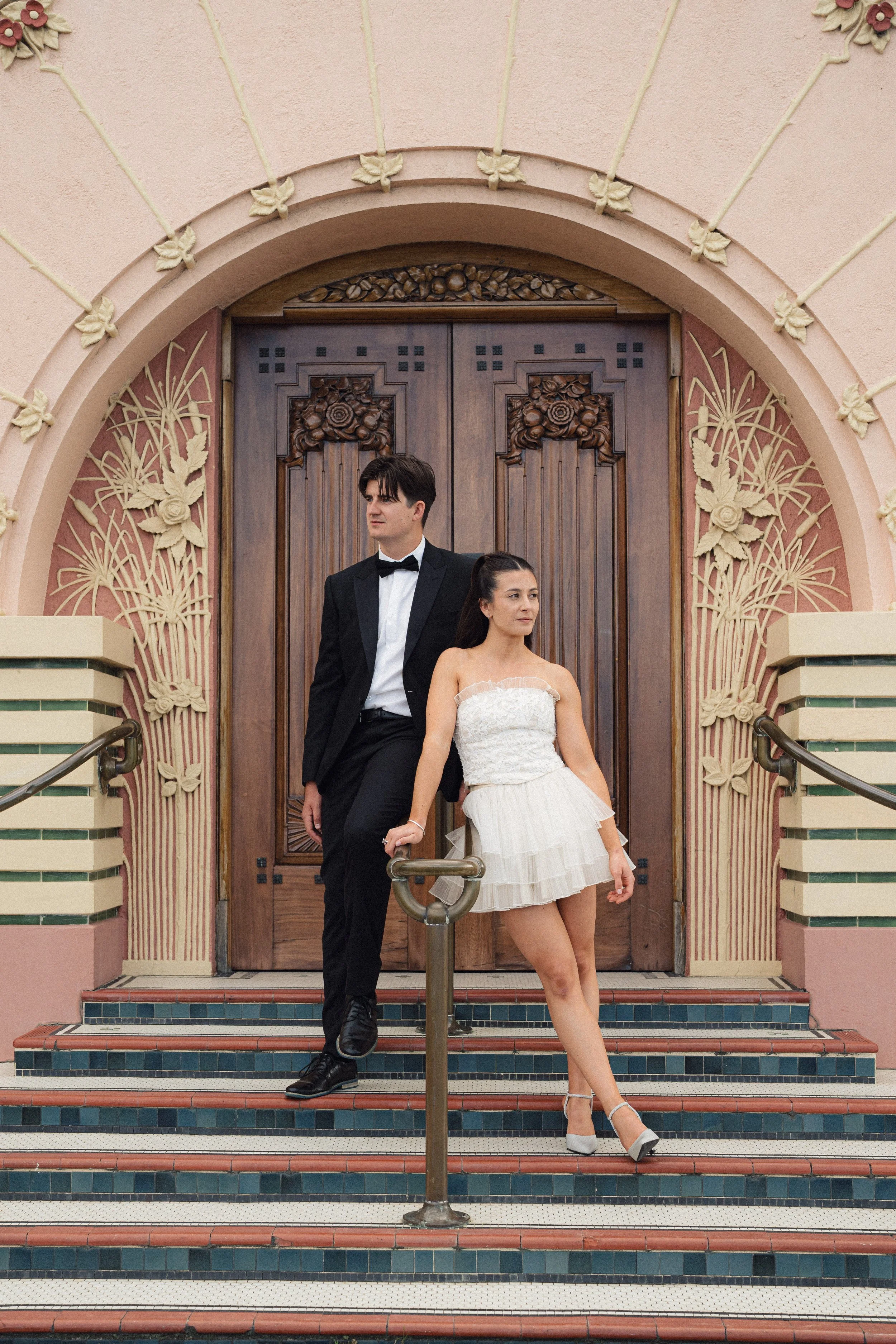 A man in a black tuxedo and woman in a white strapless dress standing on steps in front of a decorative wooden door with floral and nature designs.