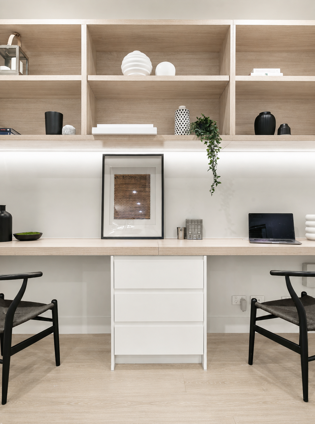 Minimalist office space with wooden shelves, white and black decorative objects, a framed artwork, a laptop, and two black chairs.