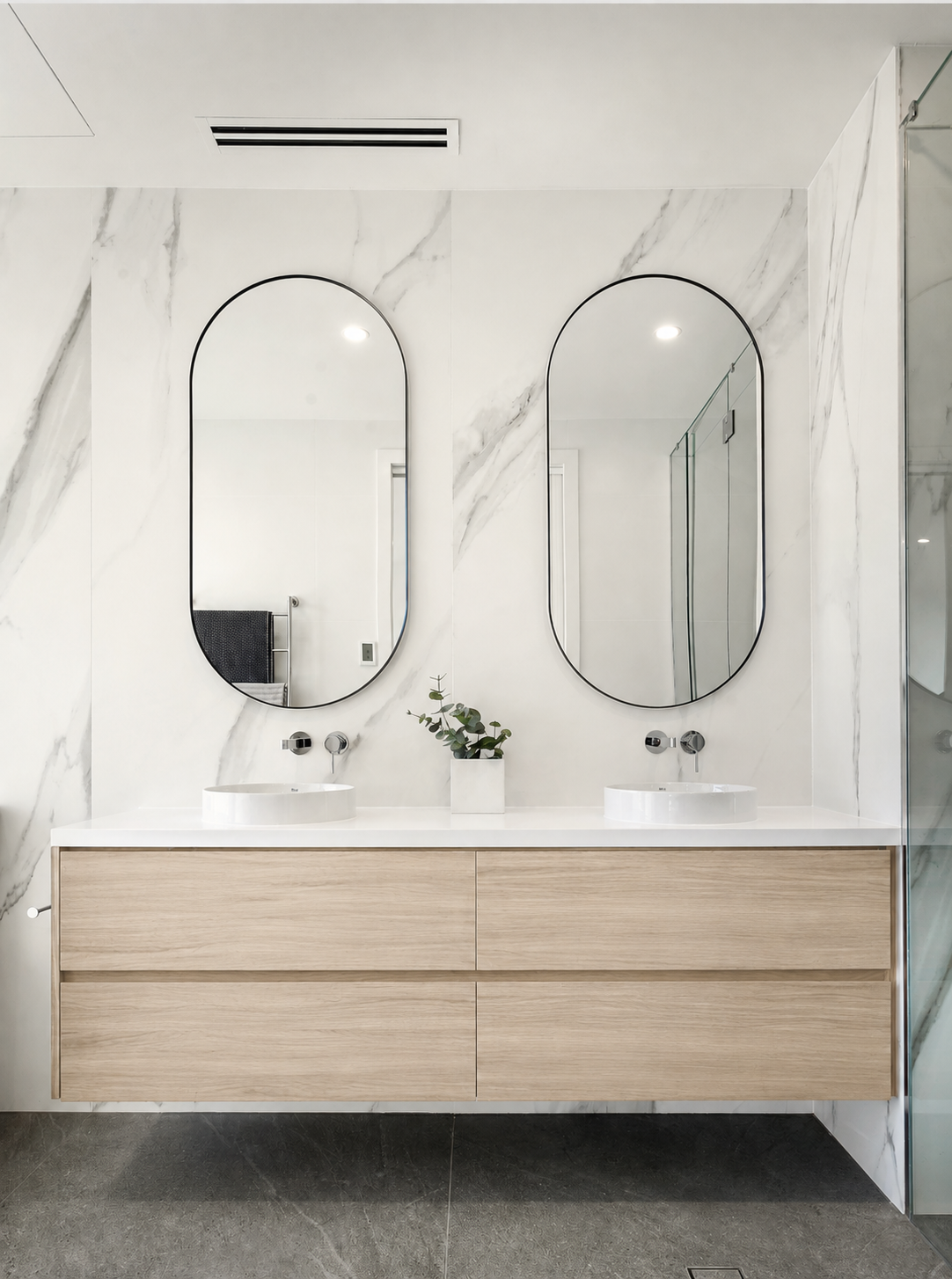 Modern bathroom with double vanity, two oval mirrors, white marble wall, small plant, dark flooring.
