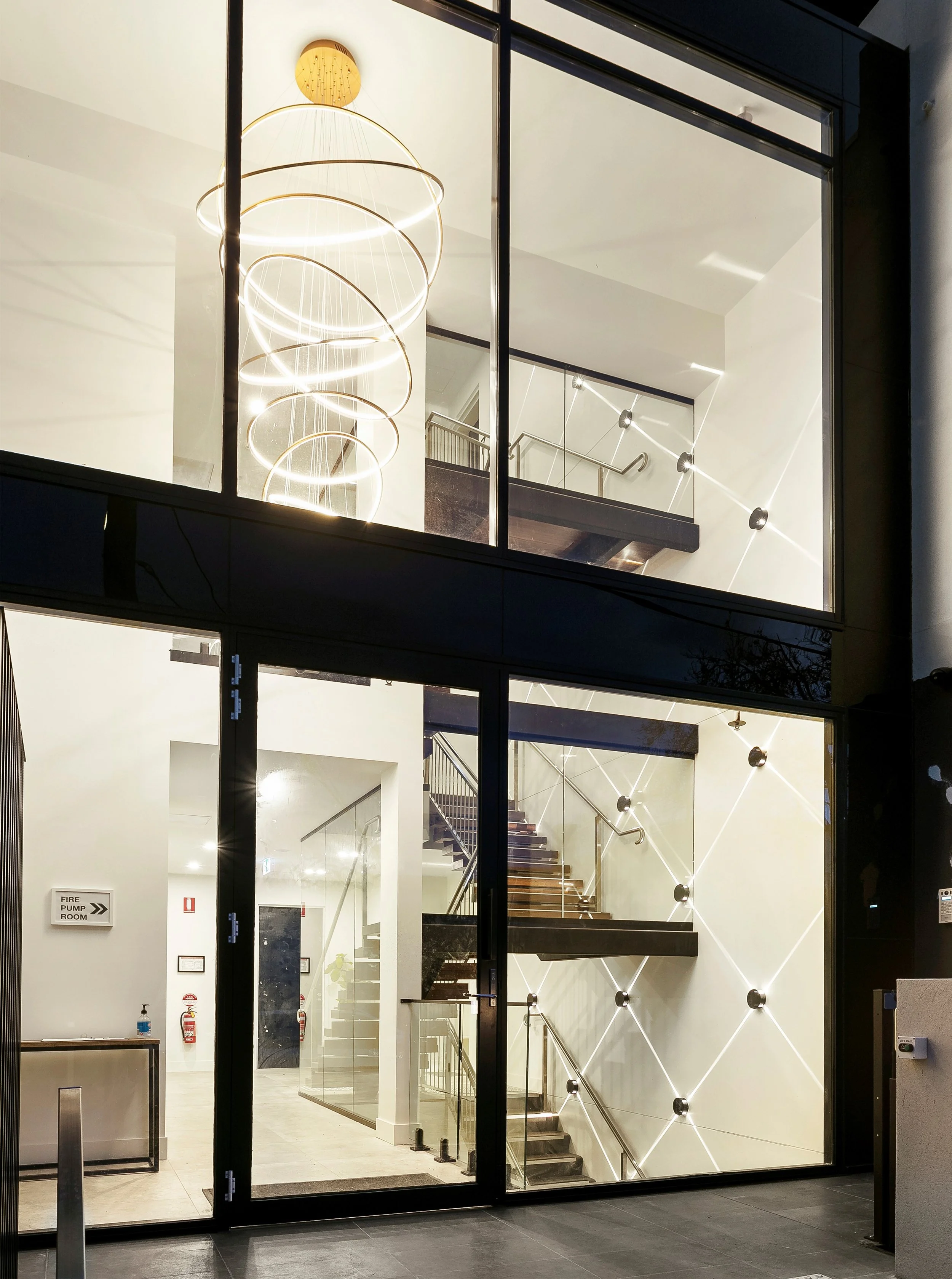 Interior view of a modern stairwell seen through a glass wall, with a spiral chandelier hanging from the ceiling.