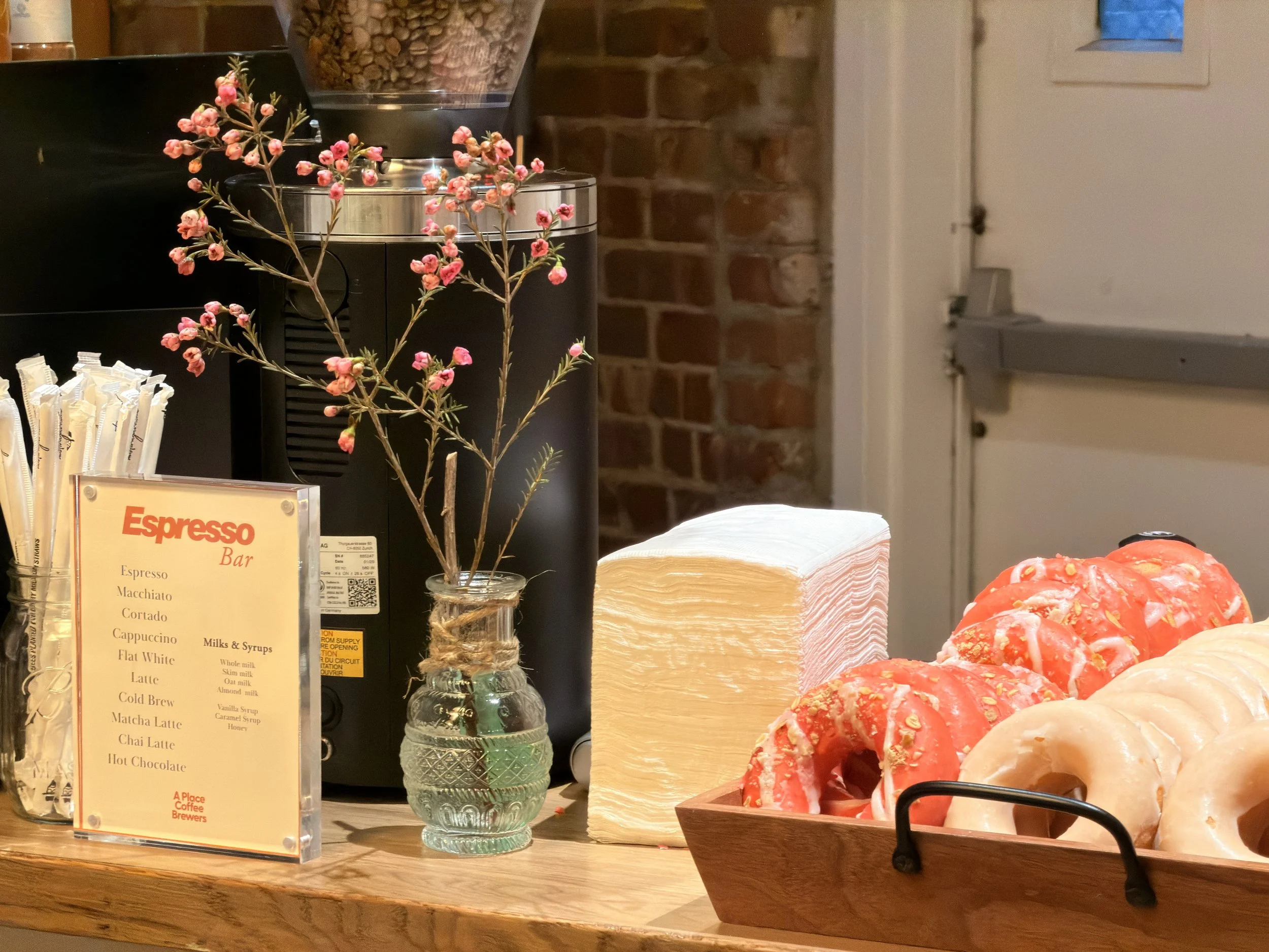 A display at a coffee shop featuring a small bouquet of pink flowers in a glass vase, a menu board listing coffee drinks, a black espresso machine, a transparent container of coffee beans, and a basket of donuts with colorful glazes and toppings, set against a brick wall background.