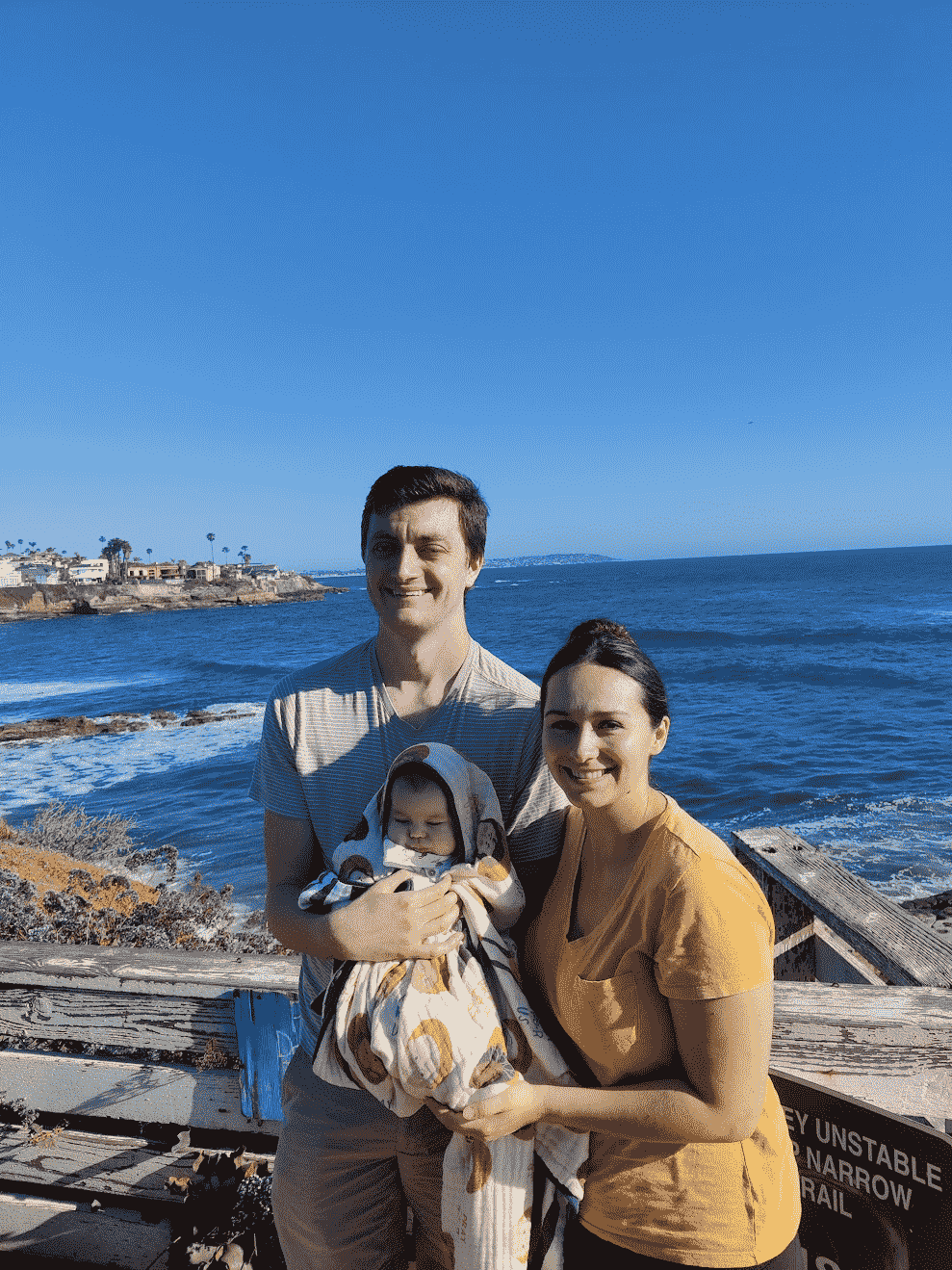New parents holding newborn in front of ocean bluffs.
