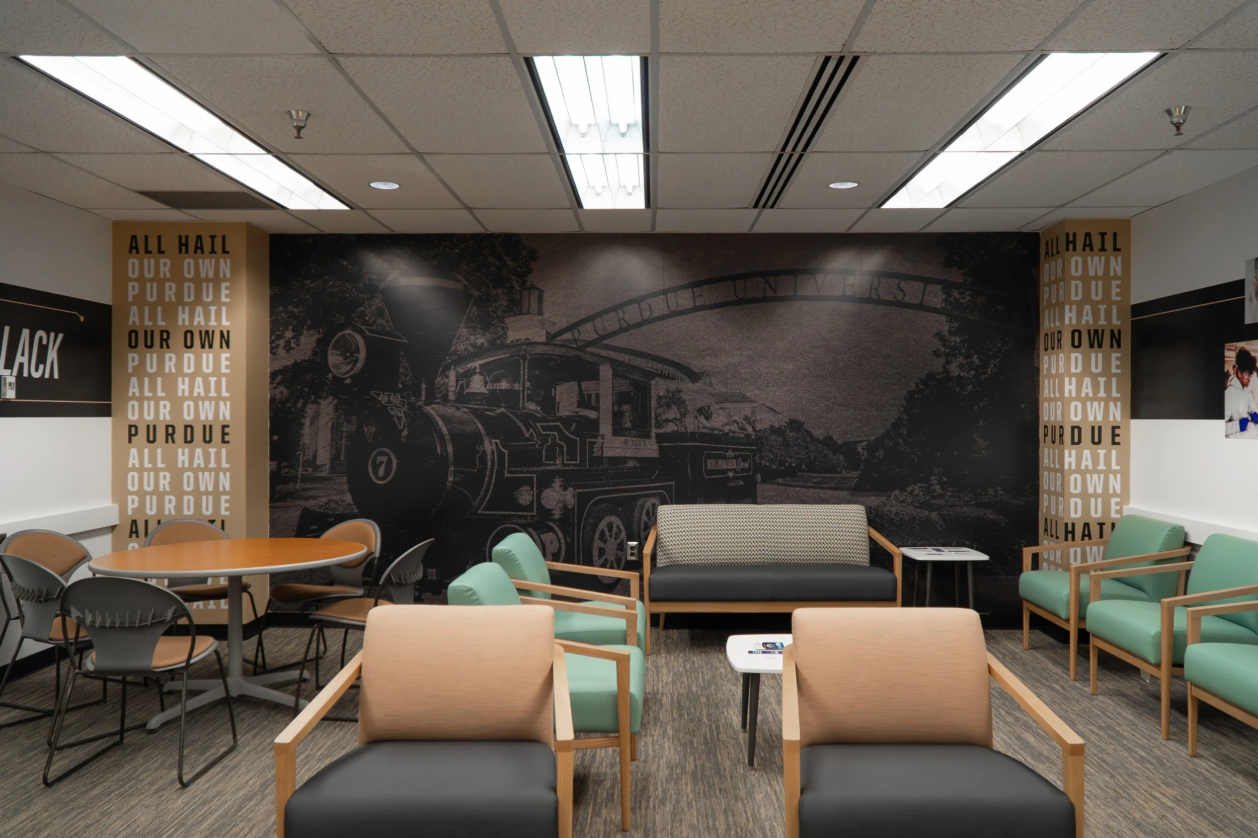 Academic advising room in Purdue University’s HAAS Hall featuring a large black-and-white wall mural of the Boilermaker Special train under the Purdue arch, flanked by gold columns with repeated “All Hail Our Own Purdue” text, and furnished with chai