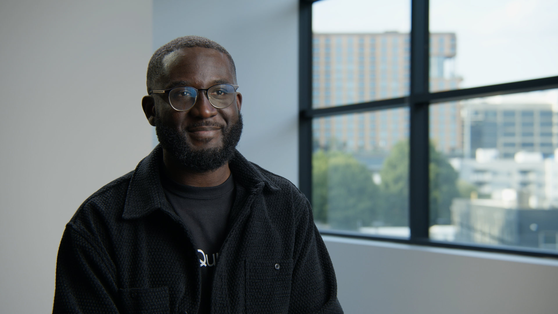 Portrait of a black man with glasses, beard, and short hair, smiling, sitting indoors near large windows with a cityscape view.