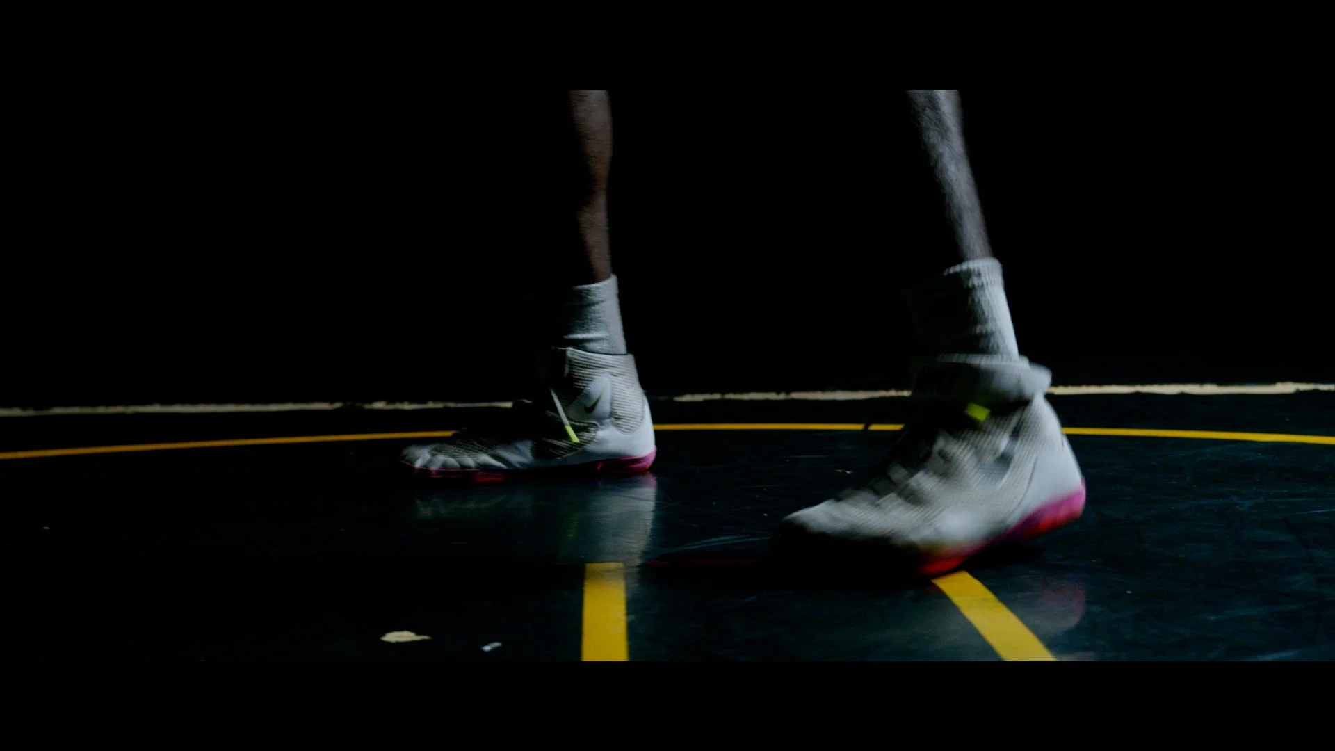 Close-up of a person wearing white Nike basketball shoes with pink soles, standing on a black court with yellow lines, dark background.