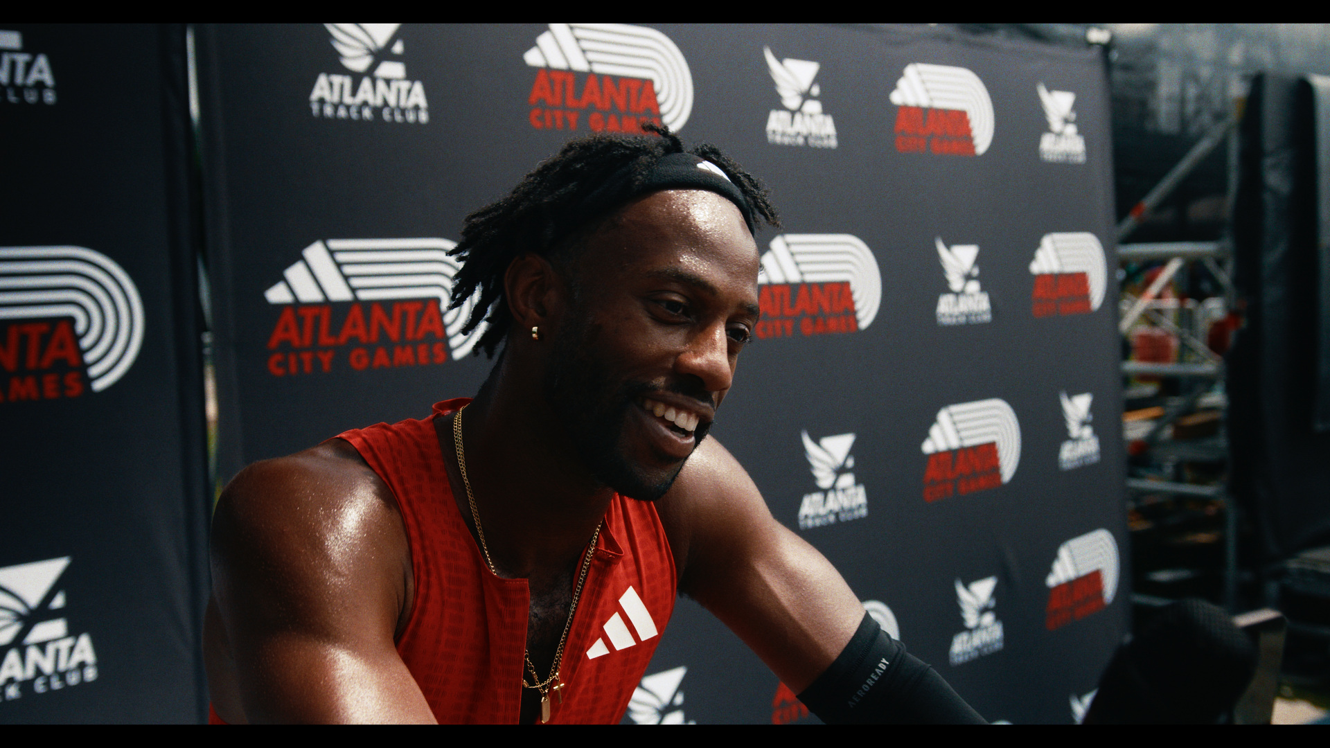 A man in athletic gear and a necklace, smiling, at the Atlanta City Games with a black backdrop featuring the event's logo.
