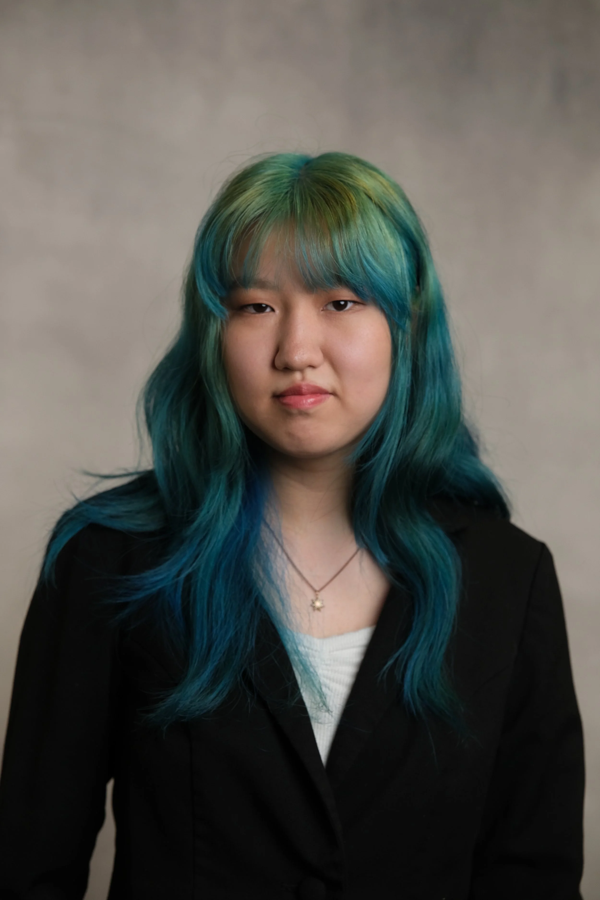 A woman with shoulder-length blue and green hair, wearing a black blazer and a white top, posing against a neutral background.