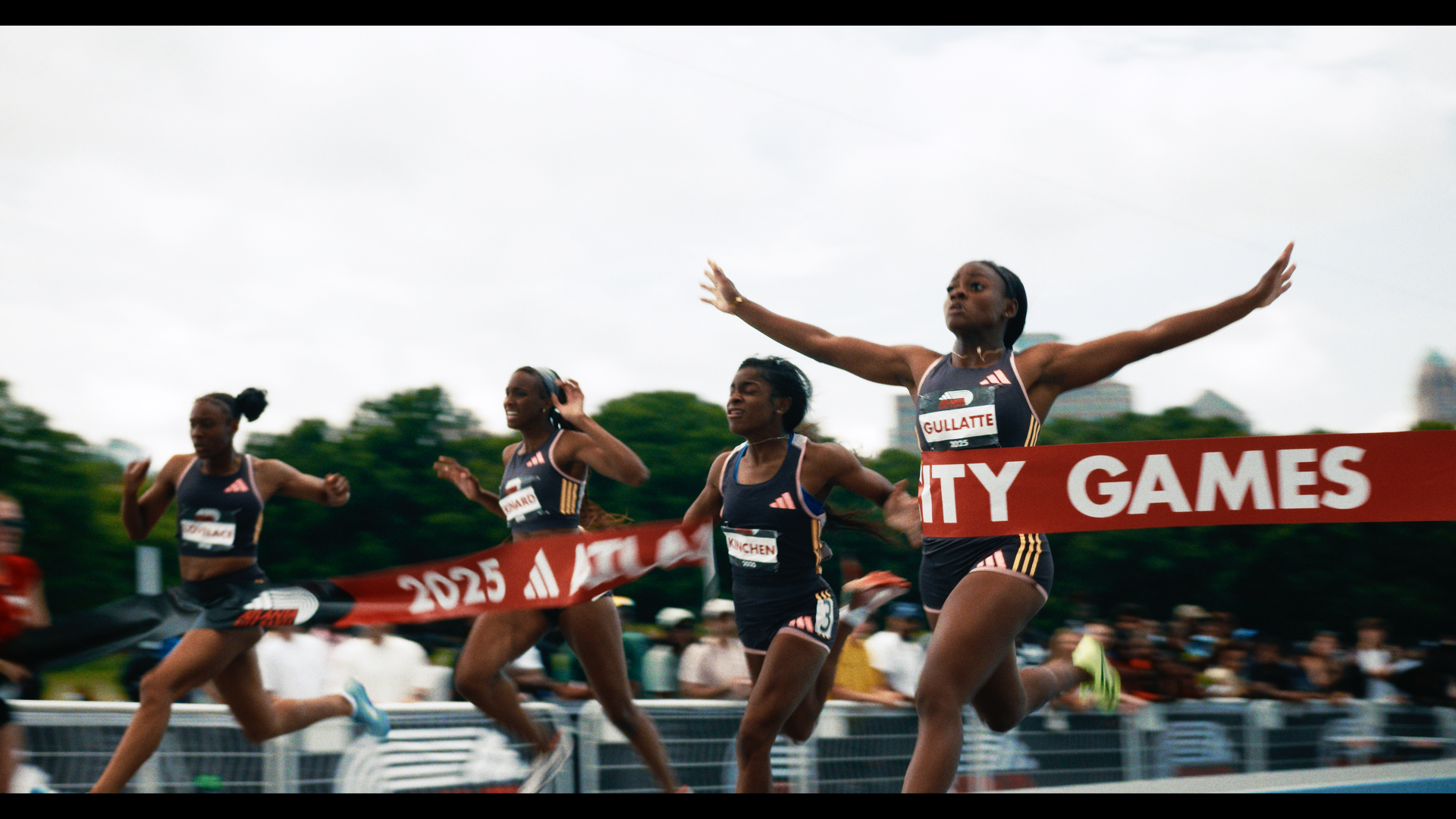 Four female athletes crossing the finish line at a race during a city games event, with one woman breaking the tape