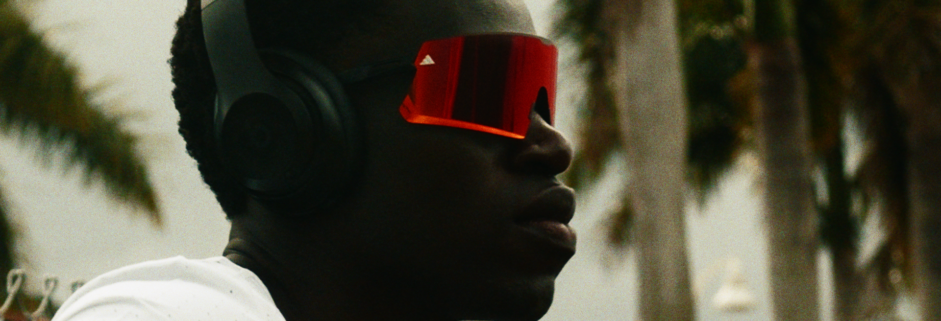 Marco Arop. Side profile of a person wearing a black cap, red reflective sunglasses, and black headphones, with a blurred outdoor background.