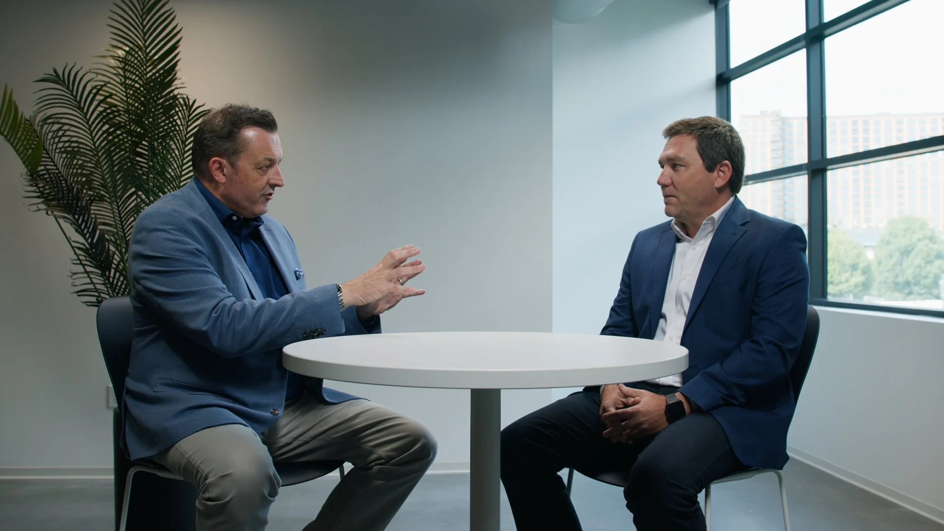 Two men in business suits sitting at a small round table in an office, engaged in conversation. There are large windows with a cityscape view behind them and a potted plant to the left.