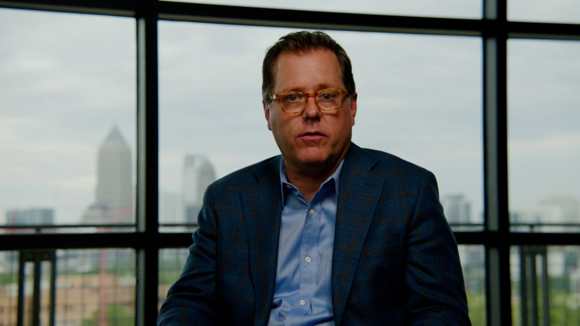 A man wearing glasses and a blue checkered blazer seated in front of large windows showing a cityscape with skyscrapers and cloudy sky.