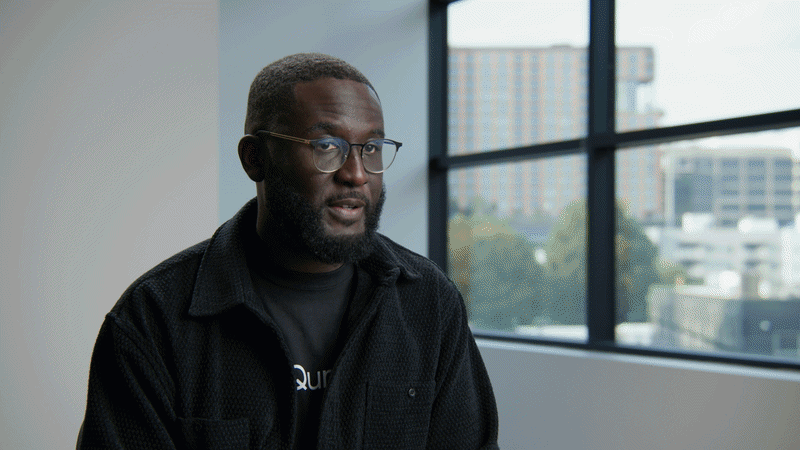 A man with glasses and a beard sitting near a window during the daytime, with a view of city buildings outside.